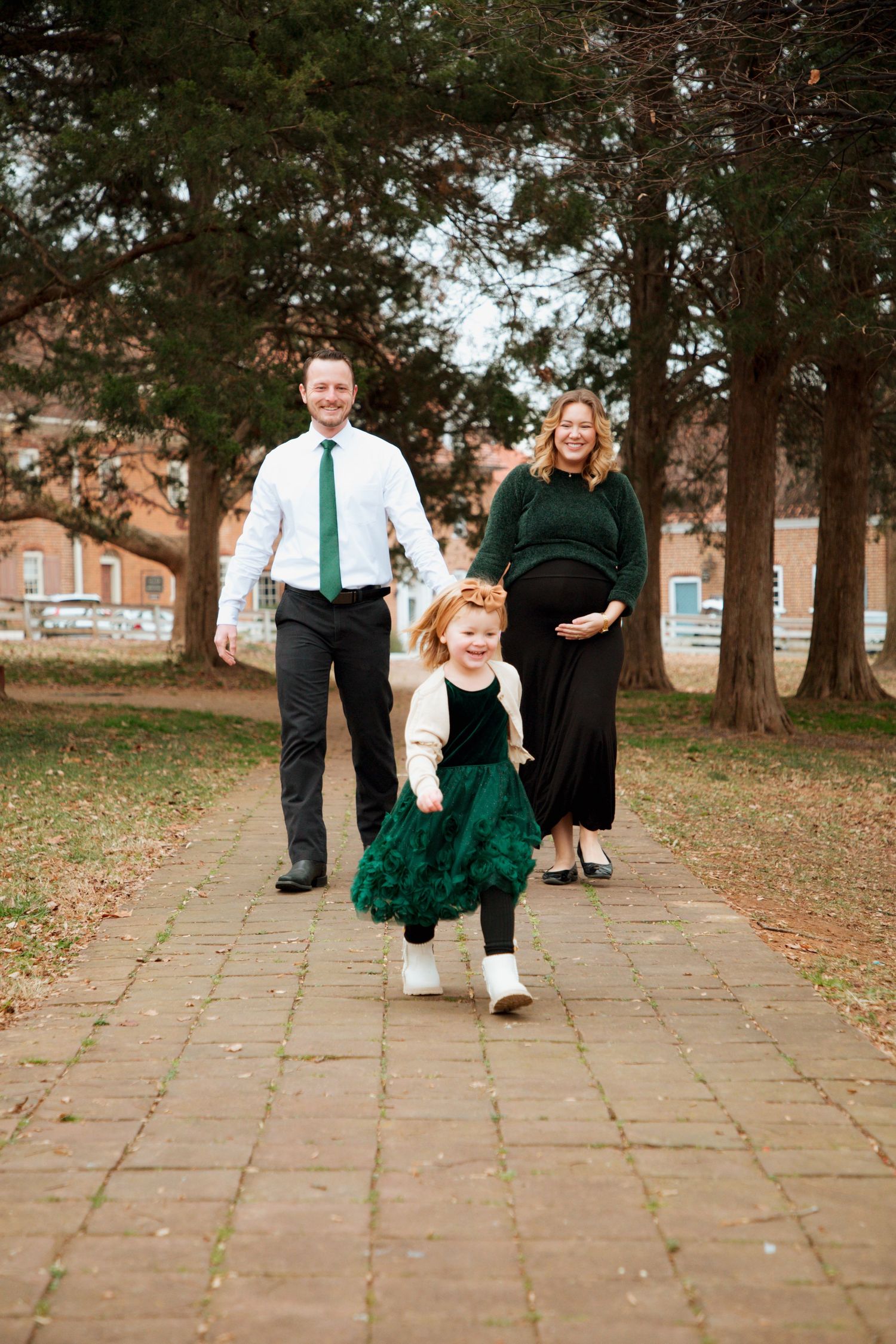 Pregnant couple smiling as toddler daughter runs toward camera during outdoor fall family session.