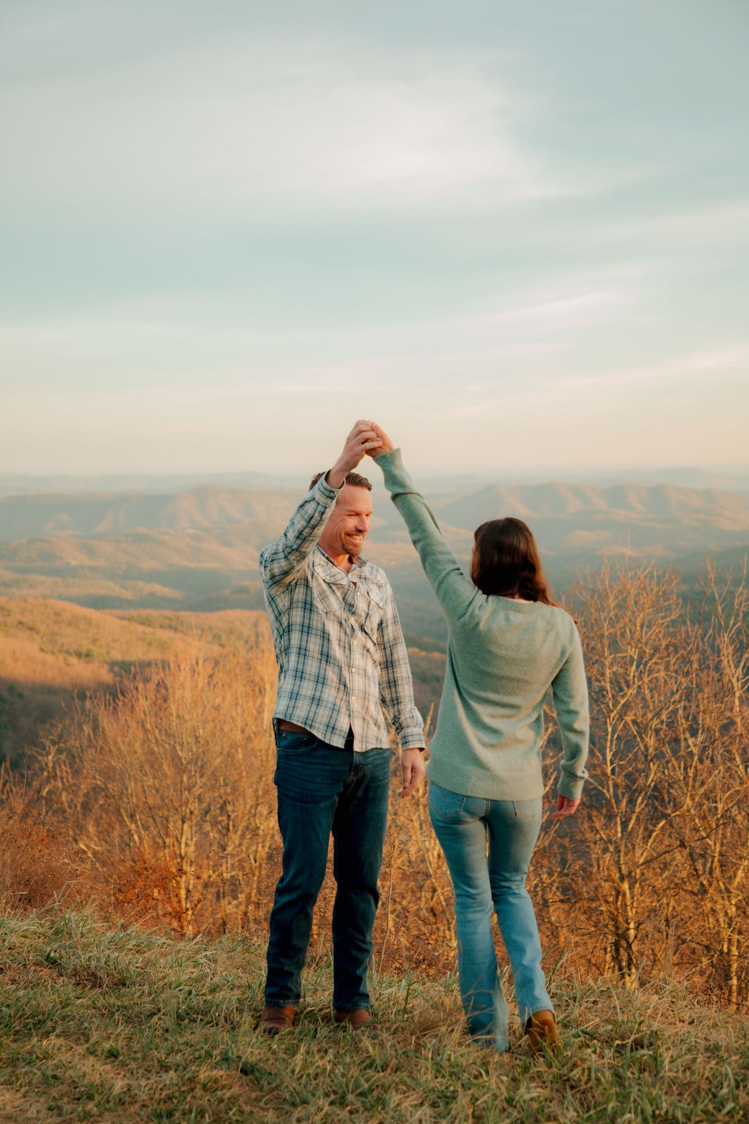 Couple dances playfully on a hilltop with expansive autumn mountain landscape behind them.