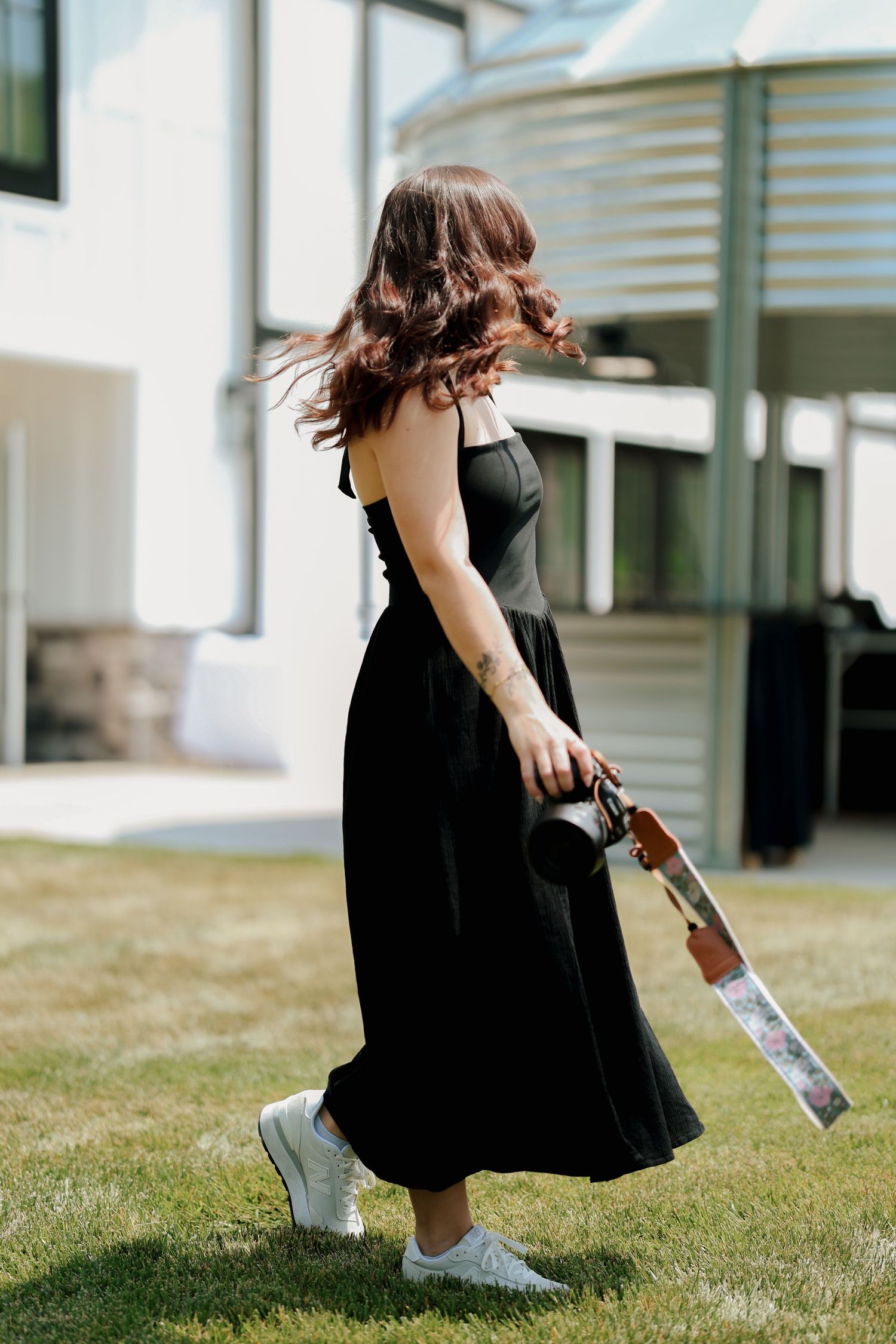 Woman in black maxi dress walks on grass holding camera with floral strap in sunlight.