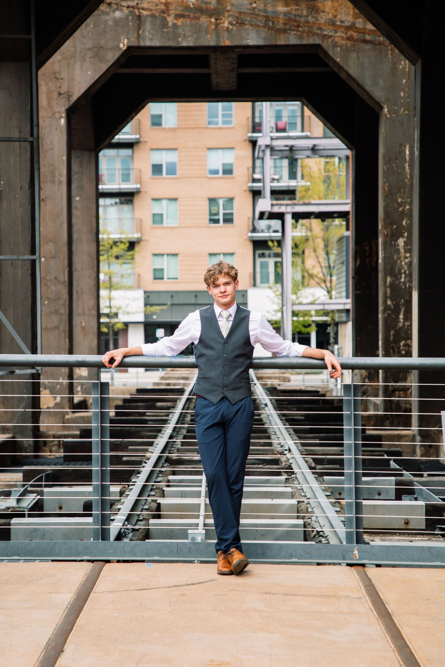 Dapper young man in navy vest and pants standing on railroad tracks under industrial bridge.