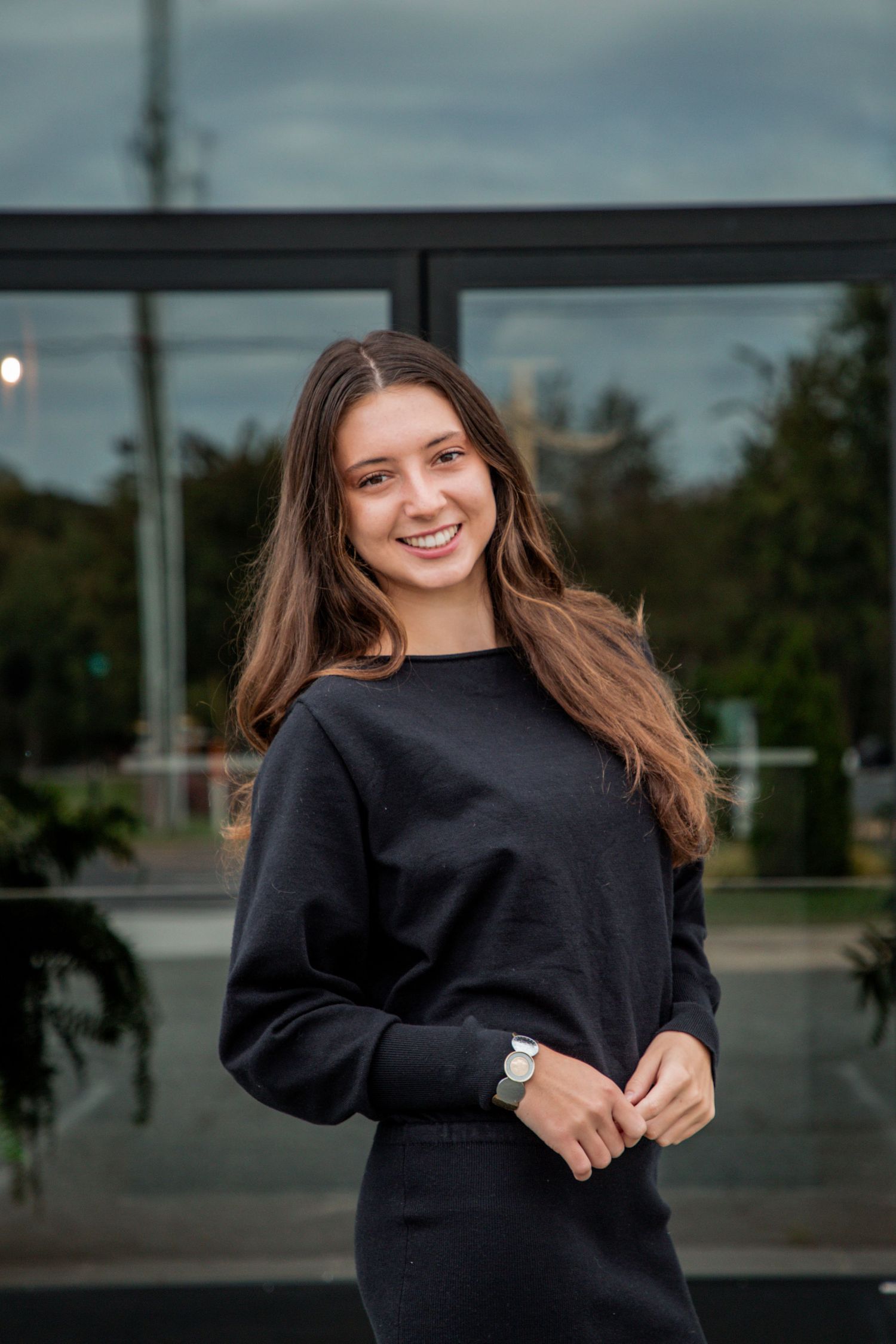 Young woman in black sweatsuit smiling confidently in front of modern glass building exterior.
