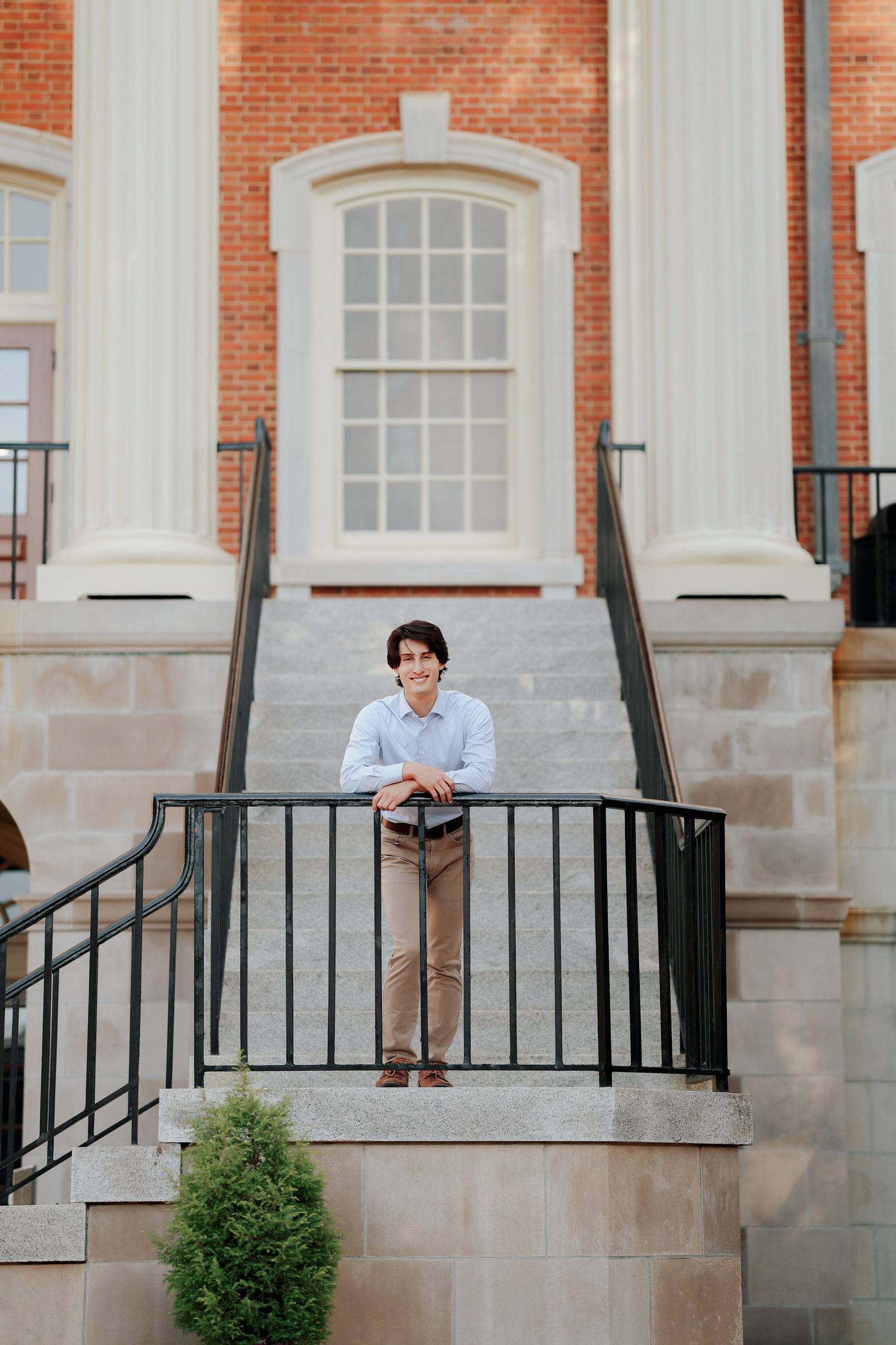 Smiling young man in blue shirt leans on railing atop stone steps of a classic brick building.