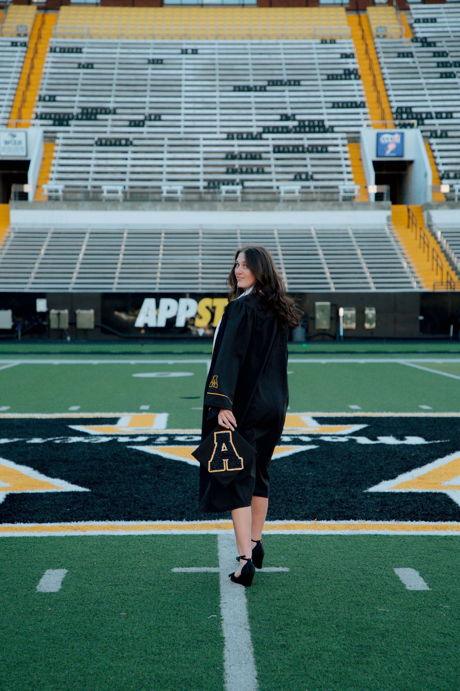 Graduate in black gown and stole posing on Appalachian State University football field at Kidd Brewer Stadium.