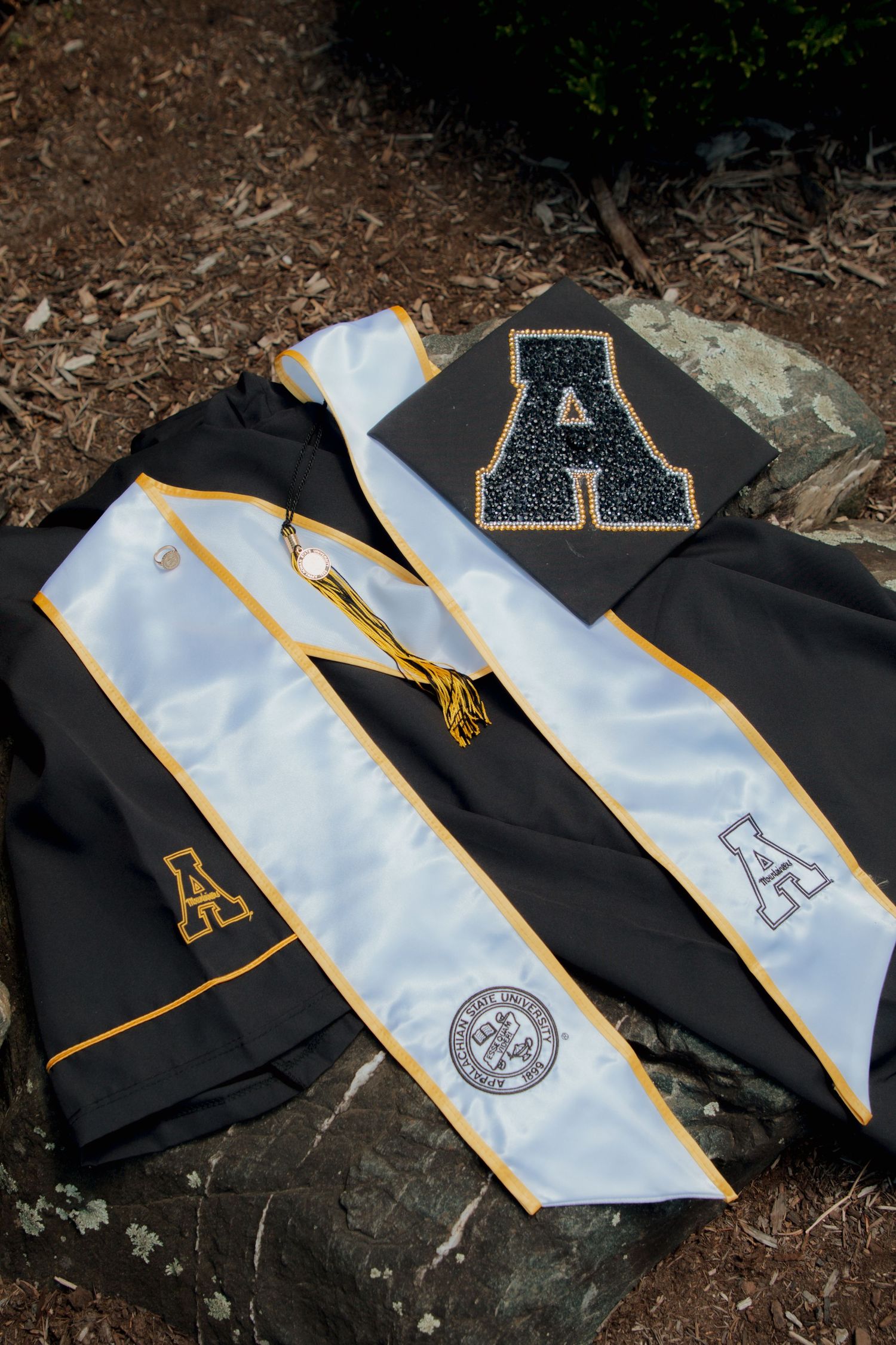 Appalachian State University graduation regalia with decorated cap, black gown, and white honor stole on rocks.
