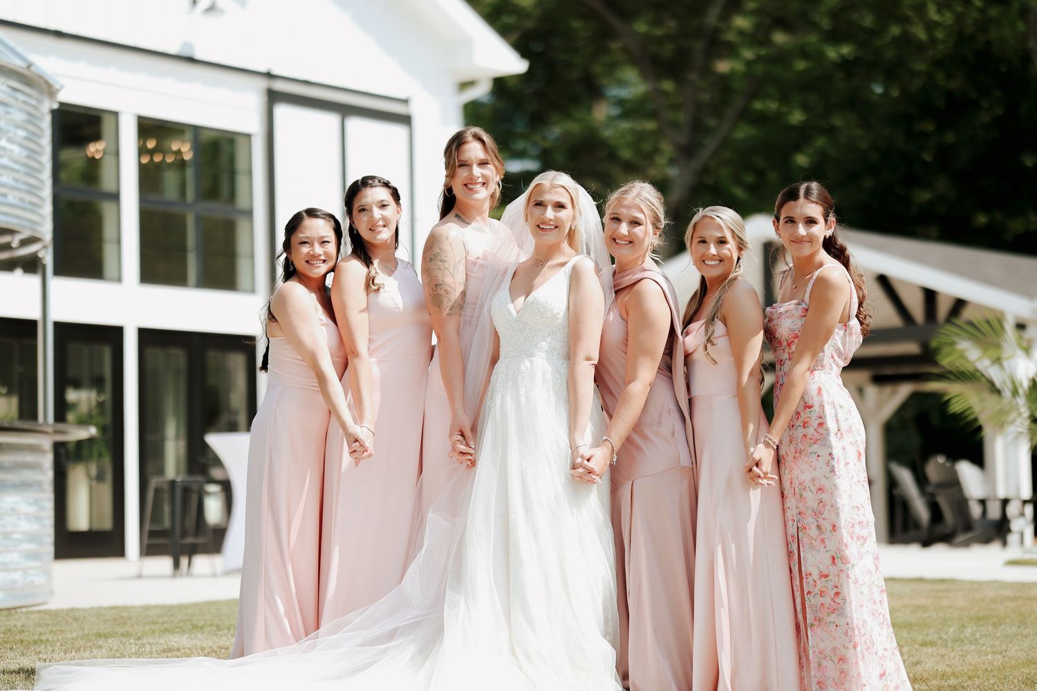 Bride in white gown posing with bridesmaids in blush dresses outside elegant wedding venue.