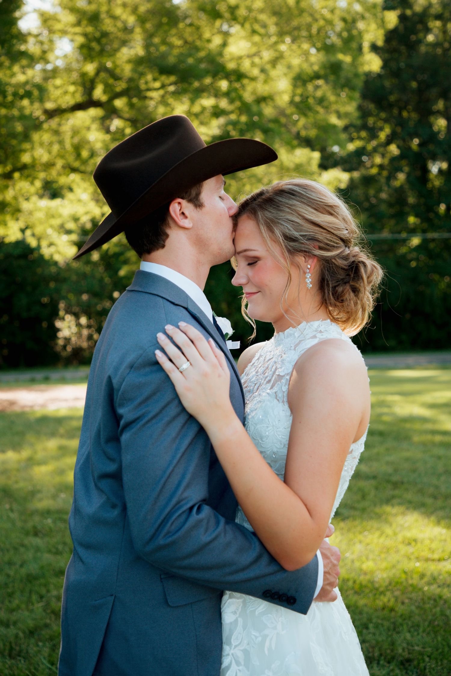 Groom in cowboy hat and blue suit kisses bride in lace wedding dress outdoors.