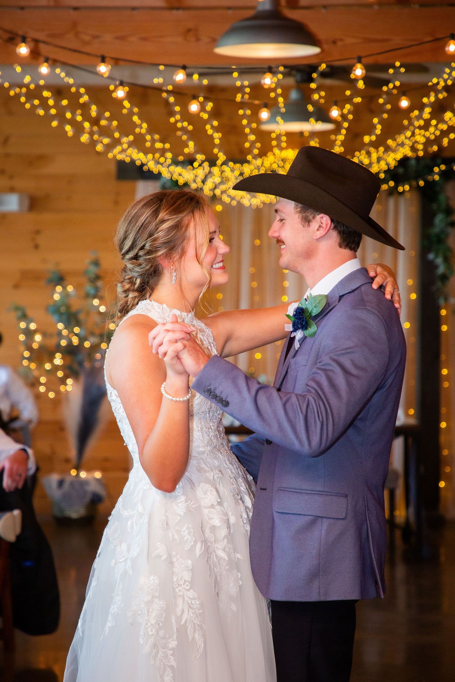 Smiling bride and groom in cowboy hat share a romantic first dance under glowing string lights at barn wedding.