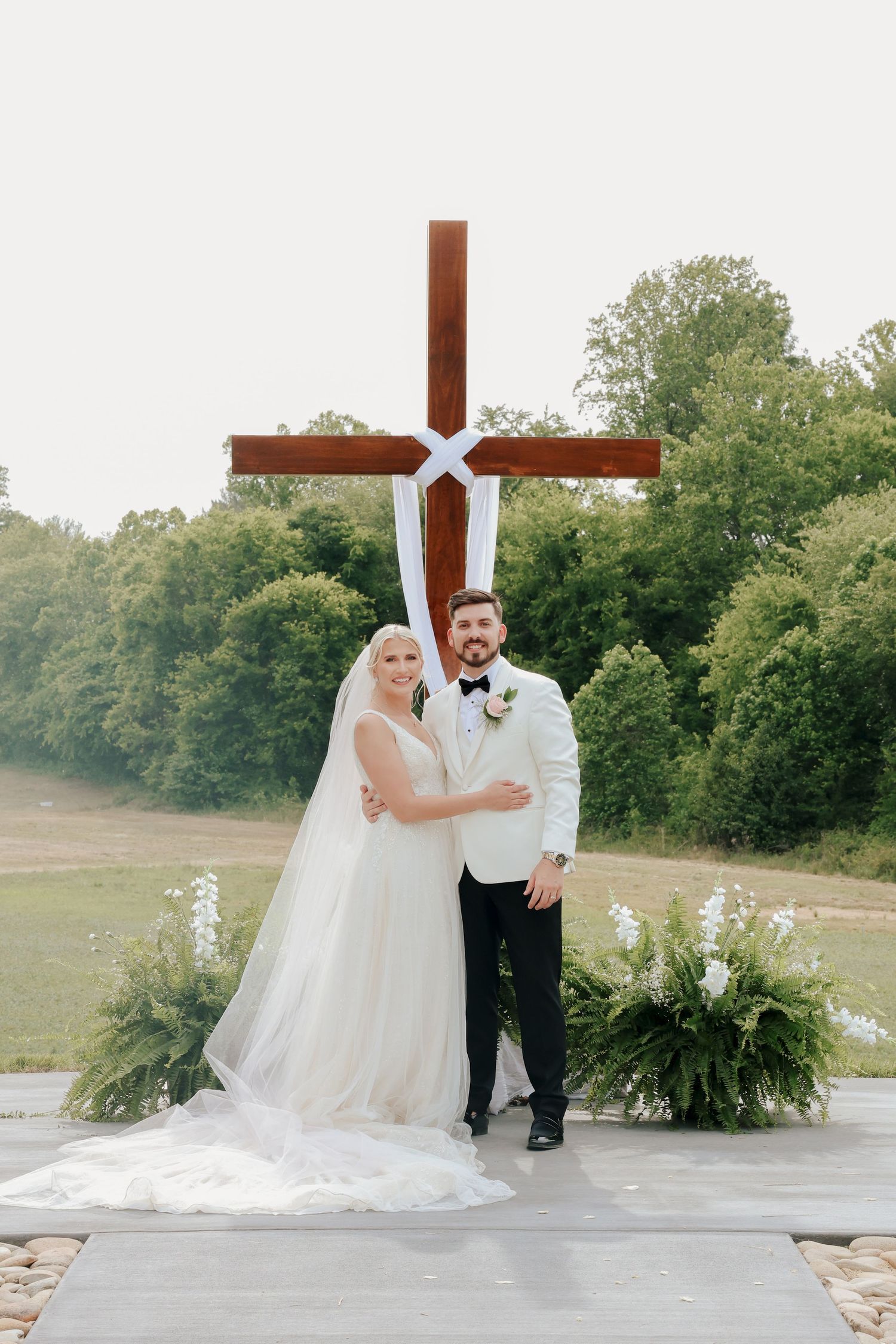 Bride and groom portrait standing before large wooden cross outdoors, bride in white gown with veil, groom in white tuxedo jacket.