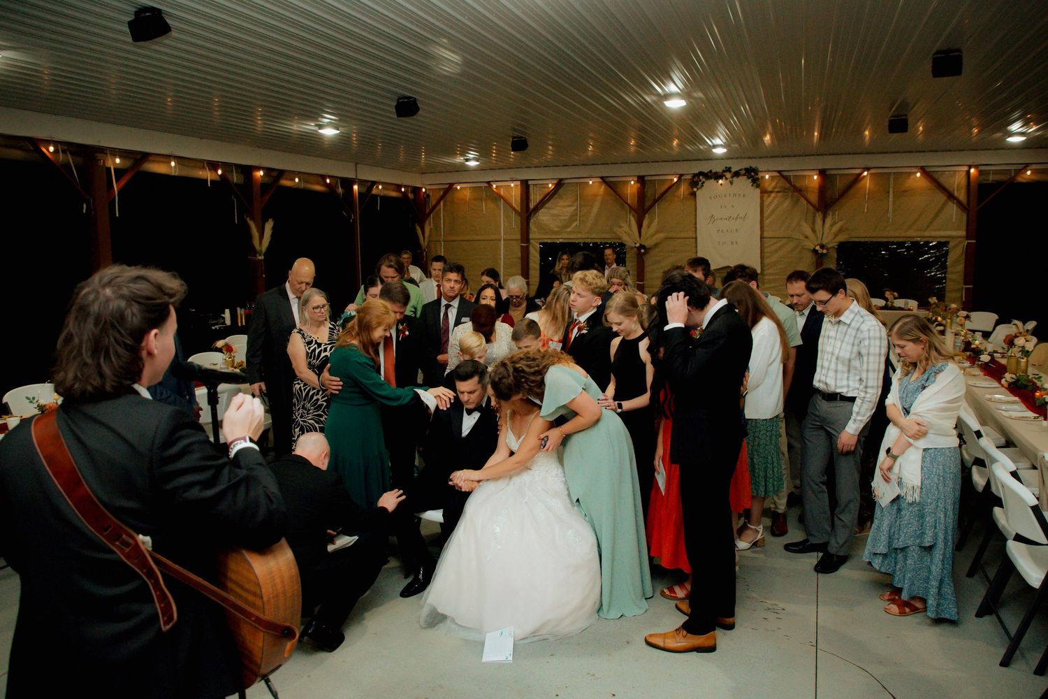 Wedding guests and bridal party gather in prayer circle inside a rustic barn reception.