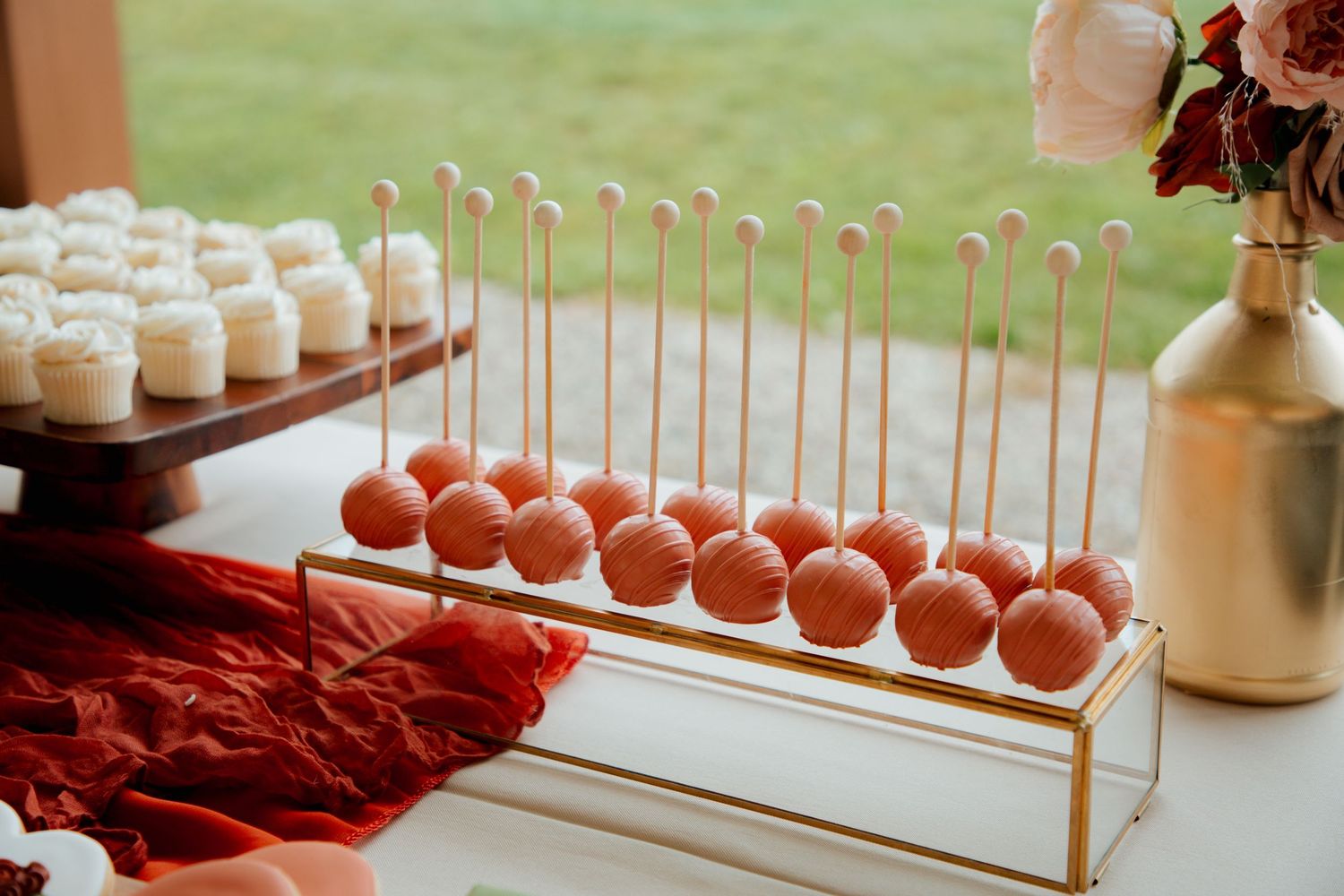 Wedding dessert table display with rows of orange cake pops on gold stand and white cupcakes on dark wood riser.