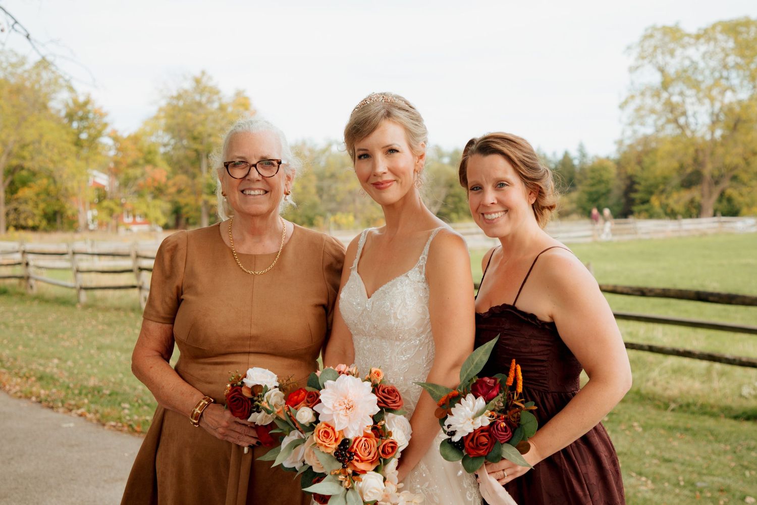 Bride with mother and bridesmaid holding fall floral bouquets outdoors.