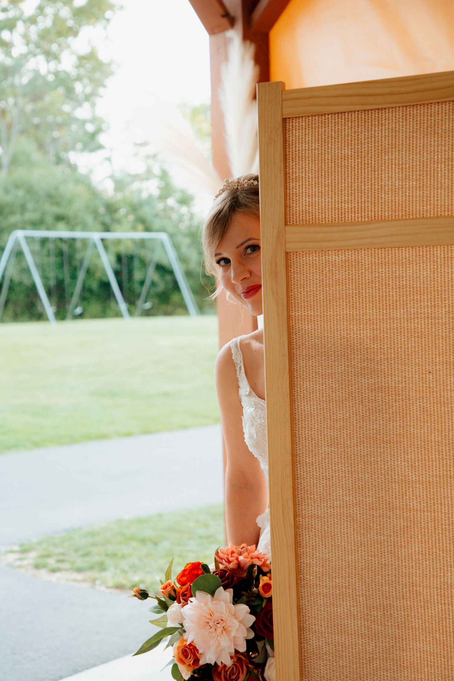 Bride peeking around a woven divider panel holding a vibrant fall floral bouquet before ceremony.