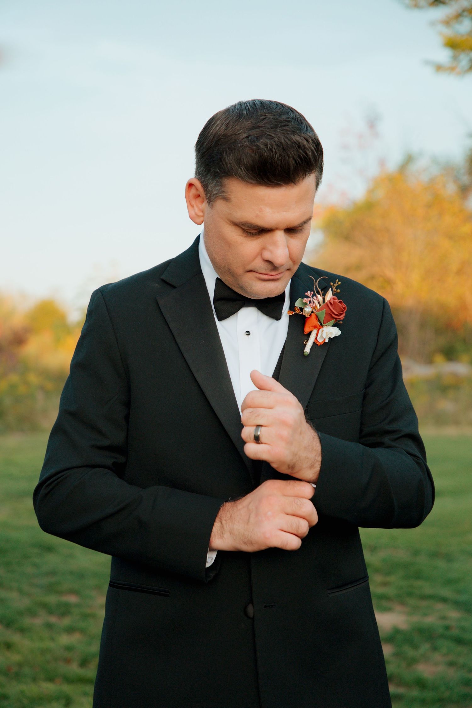 Groom in black tuxedo adjusting cufflinks with red floral boutonniere in outdoor setting.