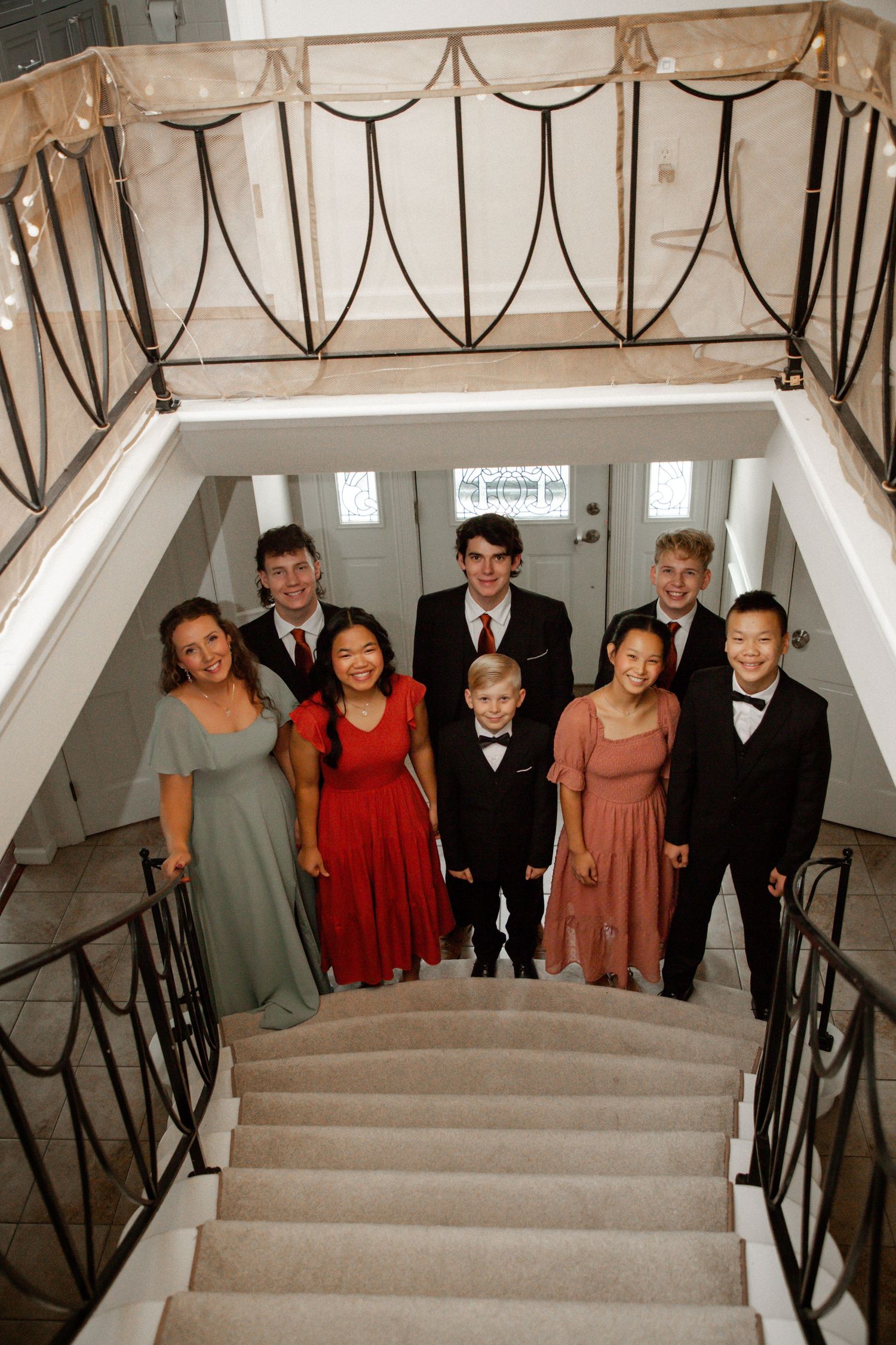 Wedding party poses on ornate staircase in formal attire including red, sage, and pink dresses with tuxedos.