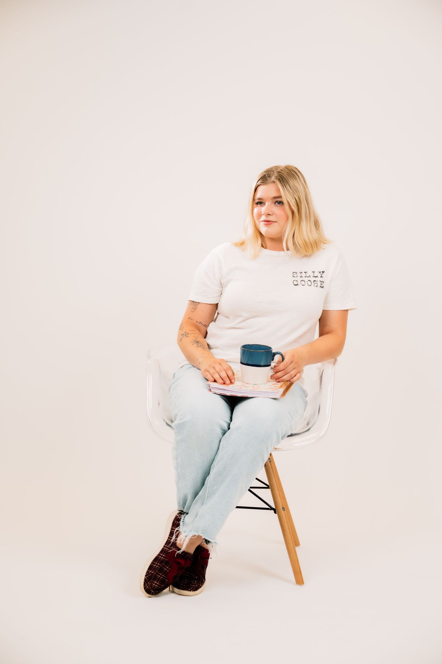 Blonde woman sitting in clear acrylic chair holding blue mug wearing white graphic tee and light wash jeans.