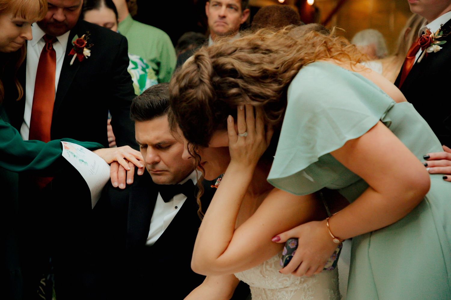 Emotional wedding moment as bride and bridesmaid embrace tearfully during reception prayer circle.