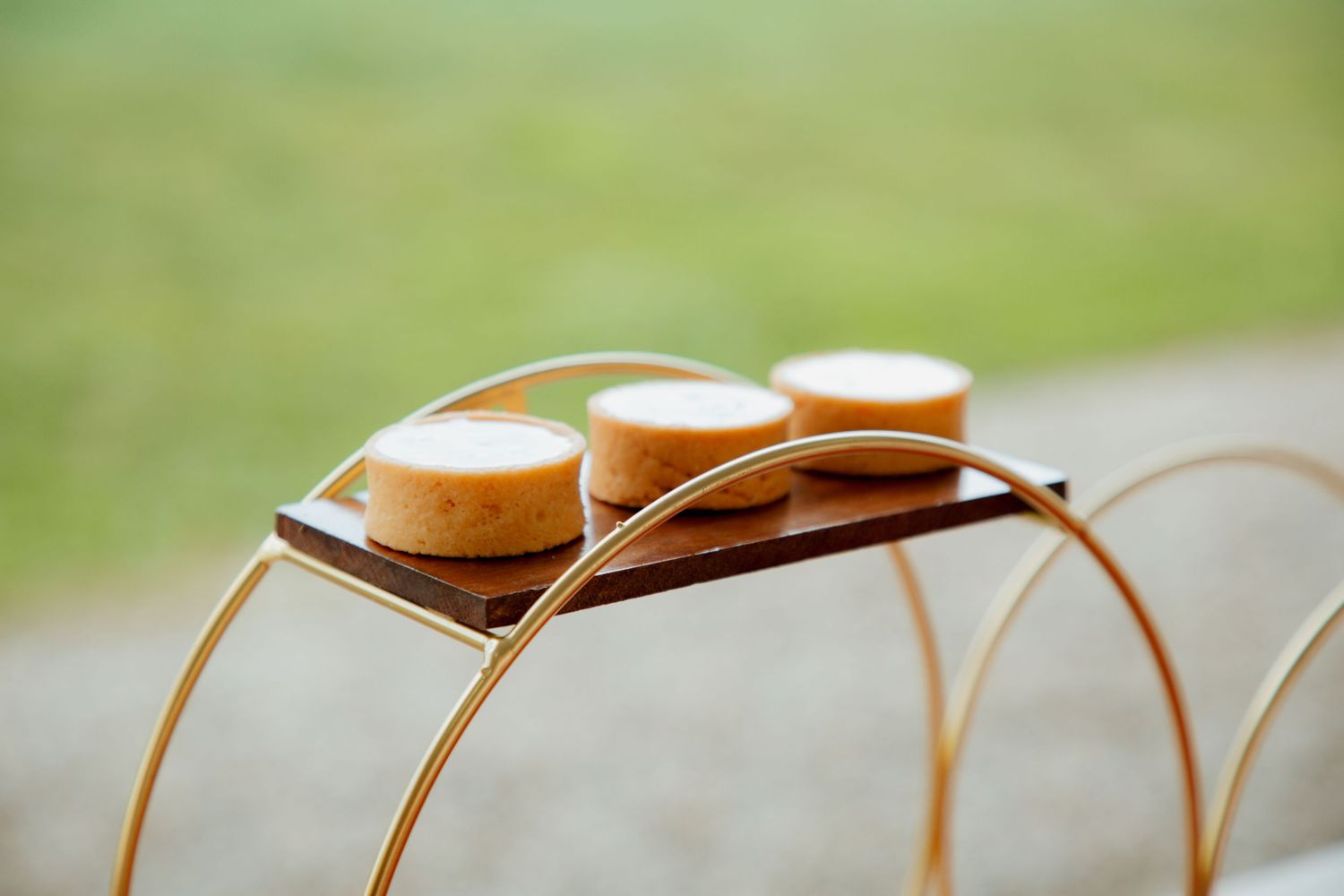 Gold wire stand holding three small round pastries on a wooden tray outdoors.