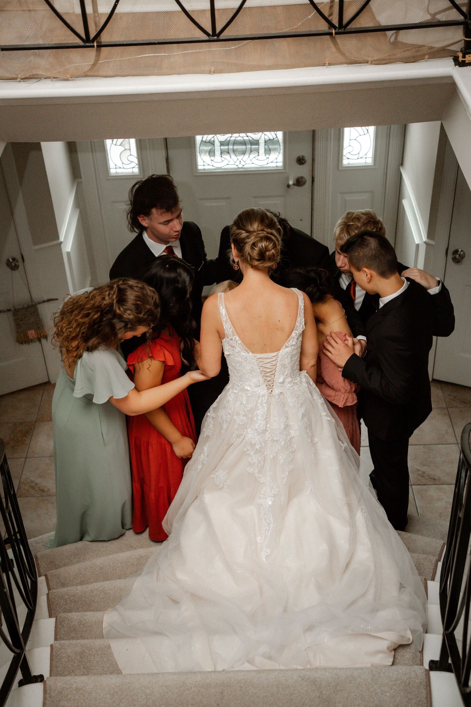 Wedding party surrounds bride in white lace gown for a group prayer on elegant interior staircase.