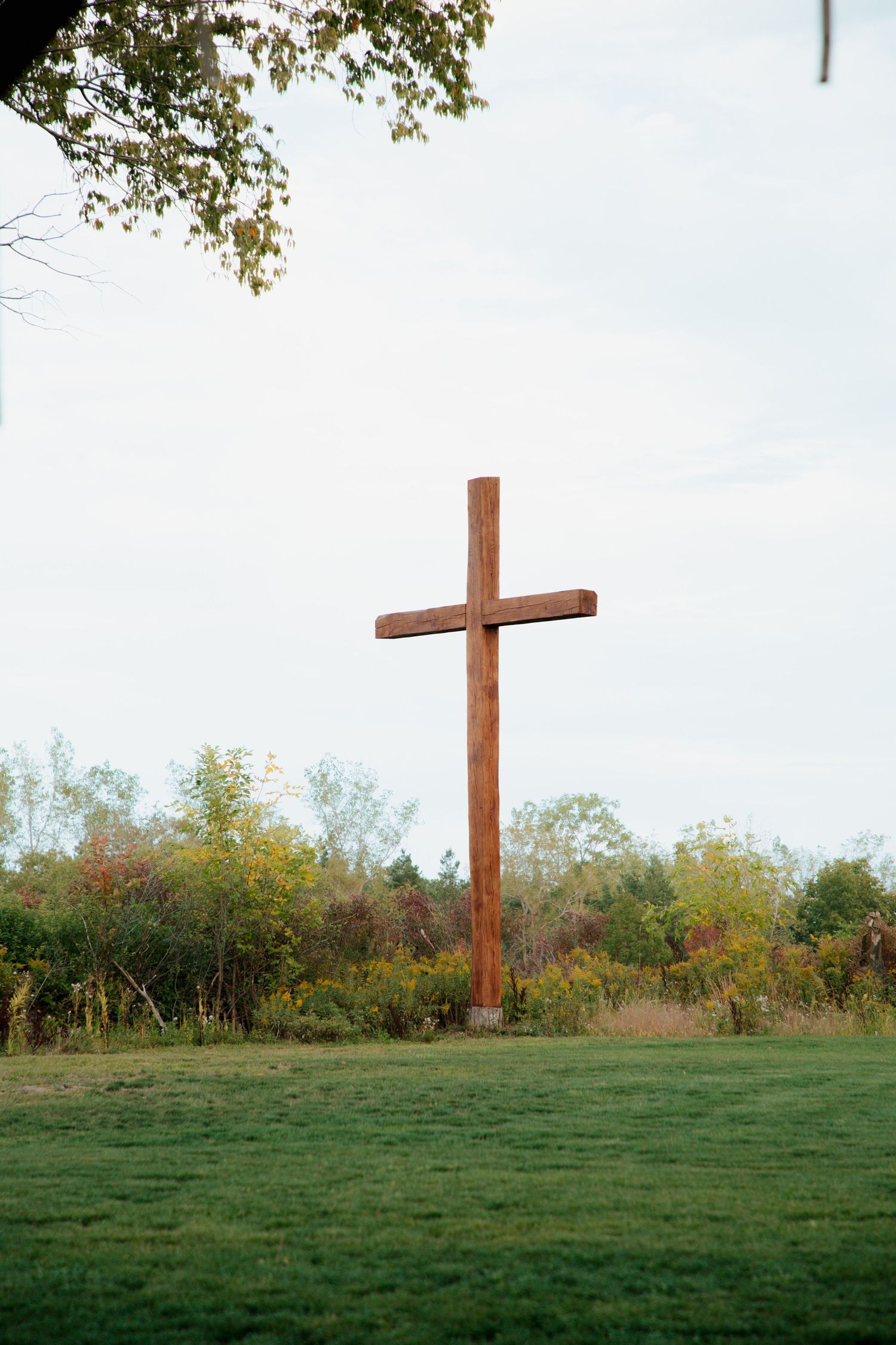 A tall wooden cross stands in a green field surrounded by colorful autumn trees under a cloudy sky.