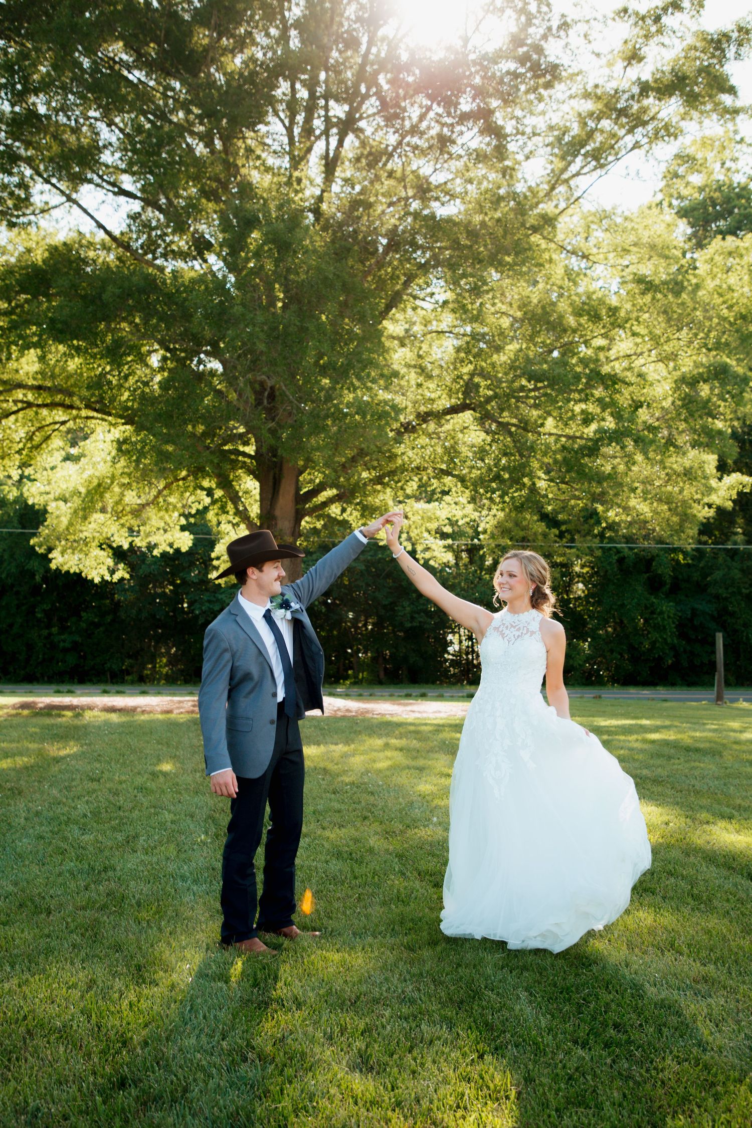 Bride and groom dance together on green lawn with sunlit trees behind them.