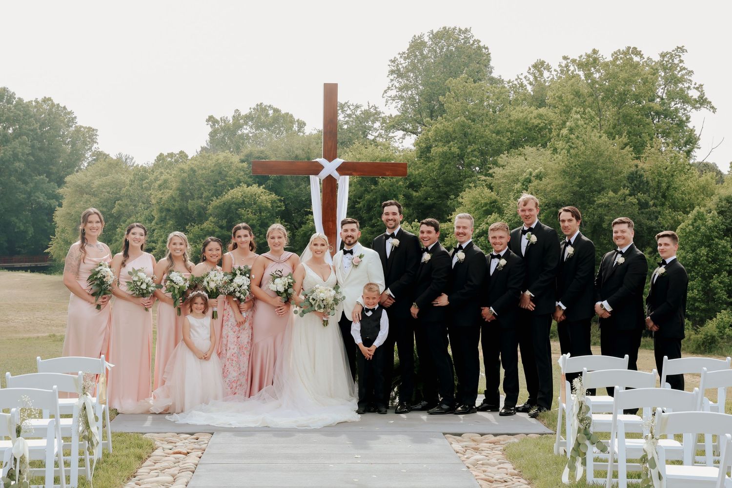 Wedding party group photo with bridesmaids in blush pink dresses and groomsmen in black suits before large wooden cross.
