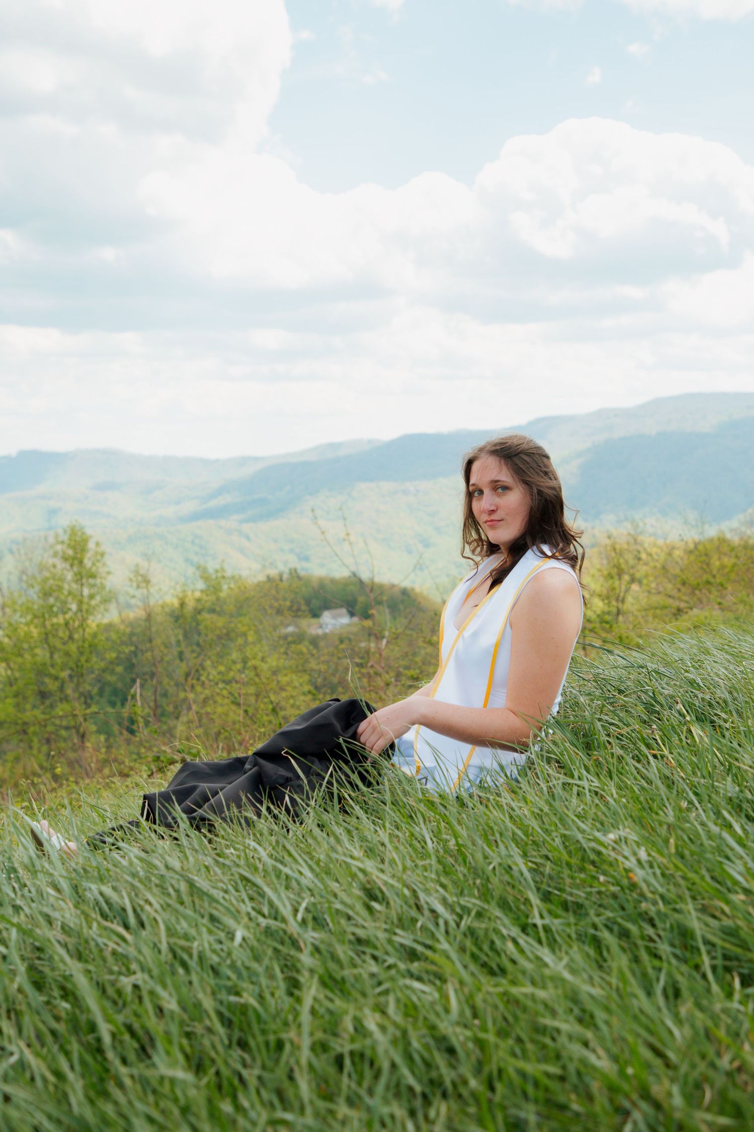 Young woman in white dress sitting in lush green grass with scenic mountain views and cloudy sky backdrop.