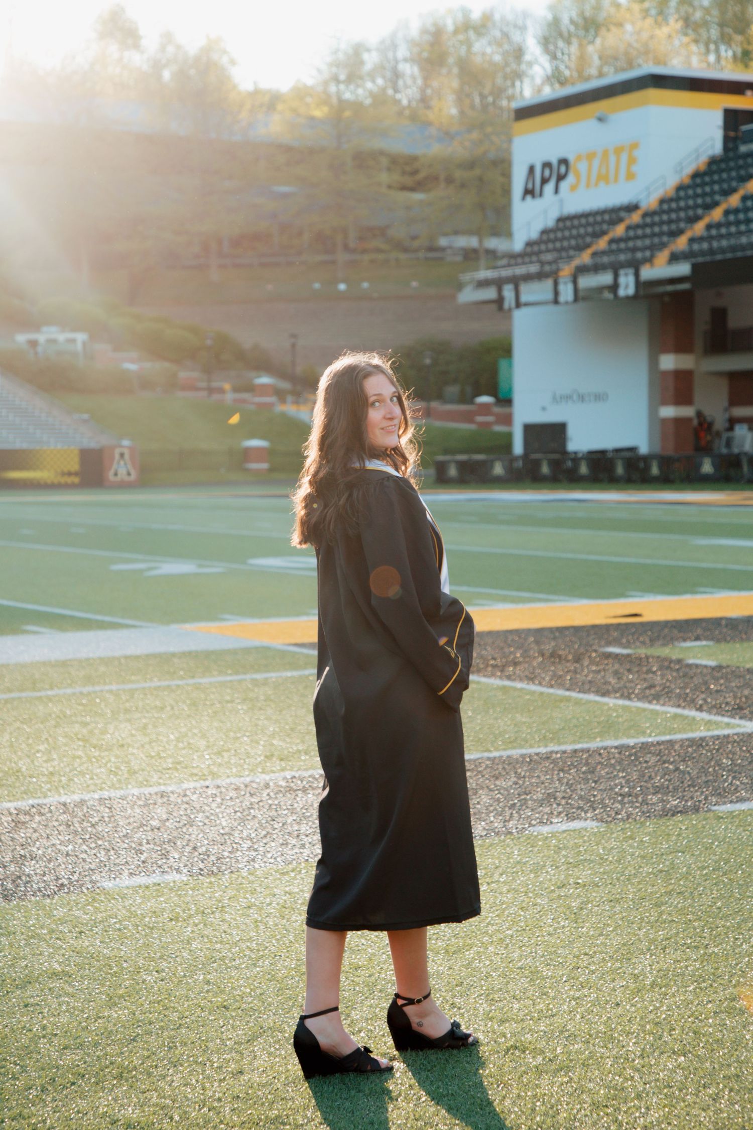Graduate in black gown poses on App State football field at golden hour sunset.