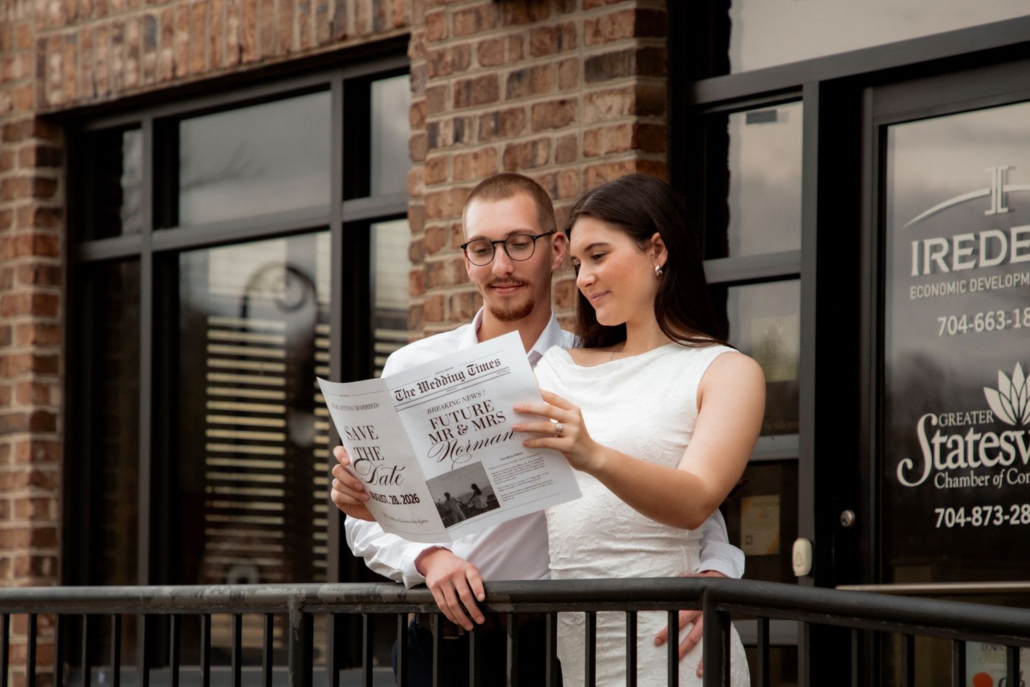 Engaged couple reads Wedding Times newspaper together on balcony outside brick building.