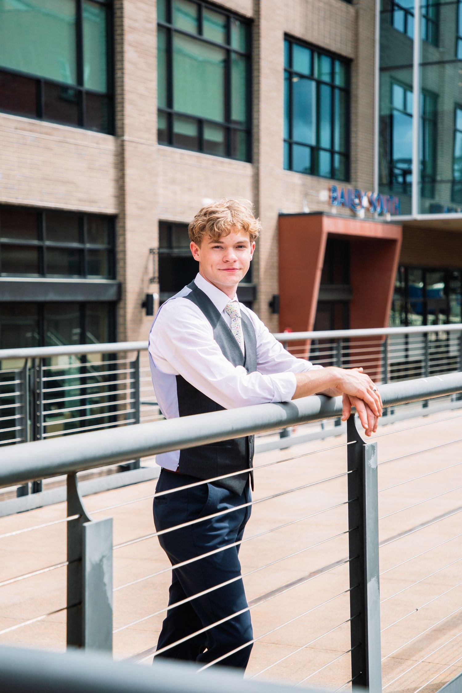 Young man in vest and tie leans on metal railing outdoors near modern brick building.
