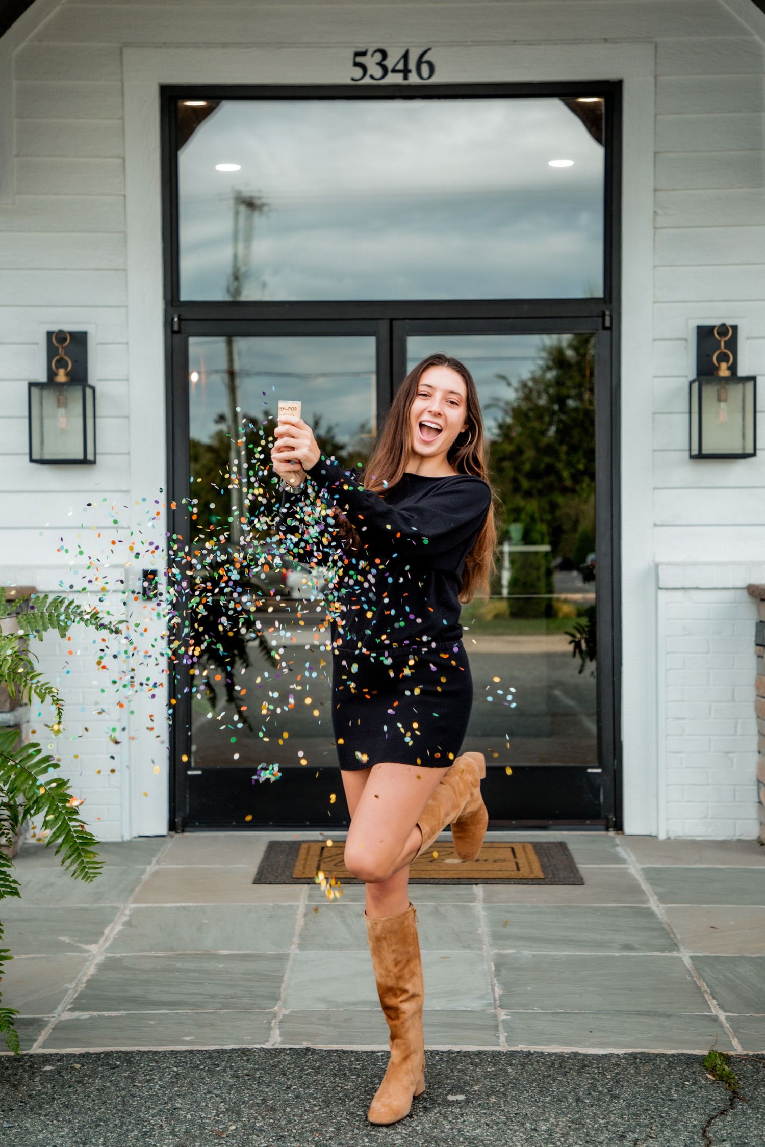 Excited young woman in black dress popping confetti cannon outside home entrance numbered 5346.