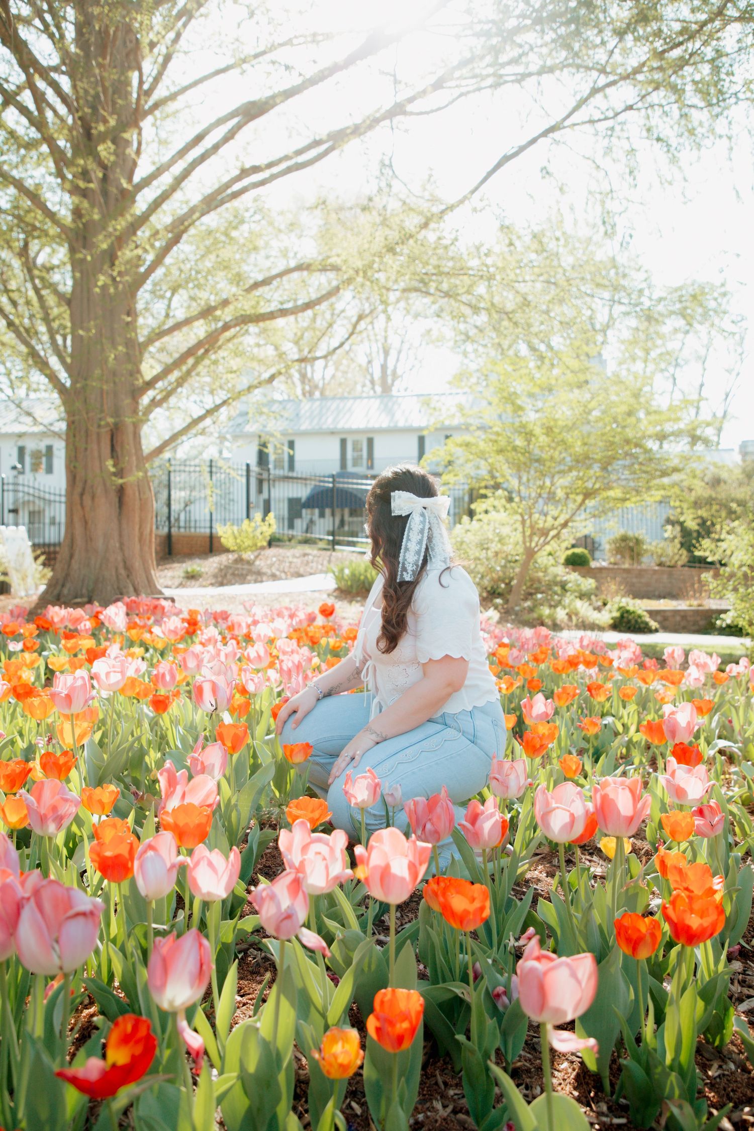 Woman with hair bow sits among vibrant pink and orange tulips in a sunny spring garden.