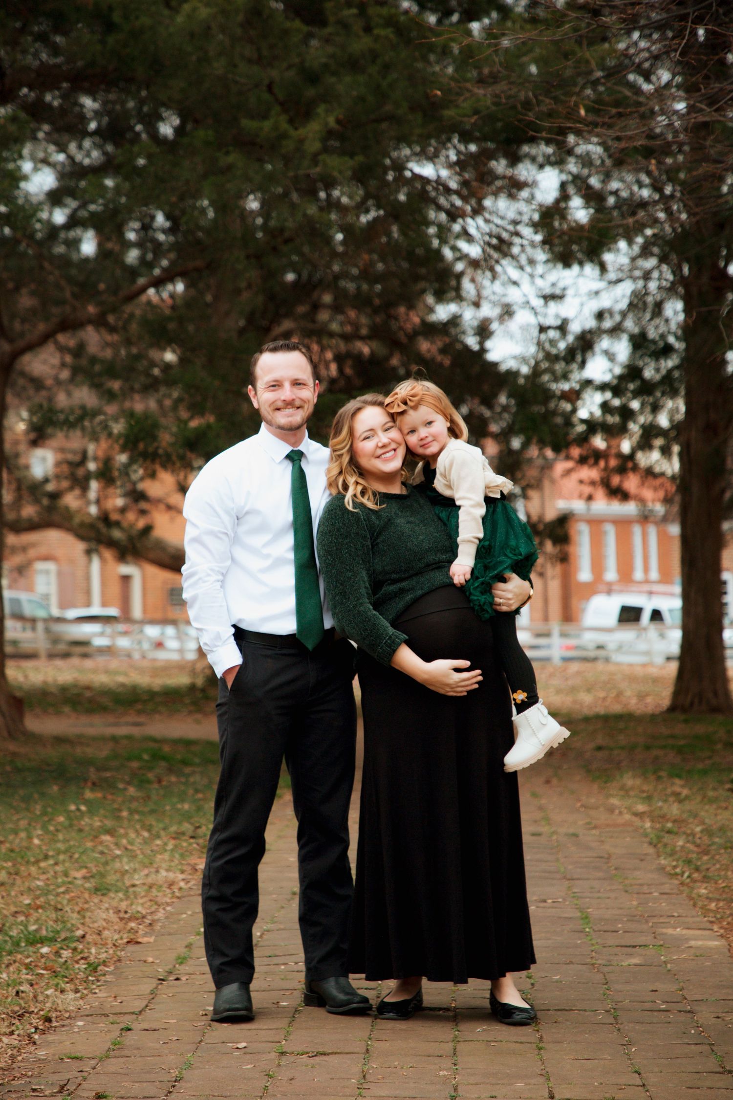 Pregnant woman in green sweater holds toddler while standing with man in white shirt and green tie on brick path.