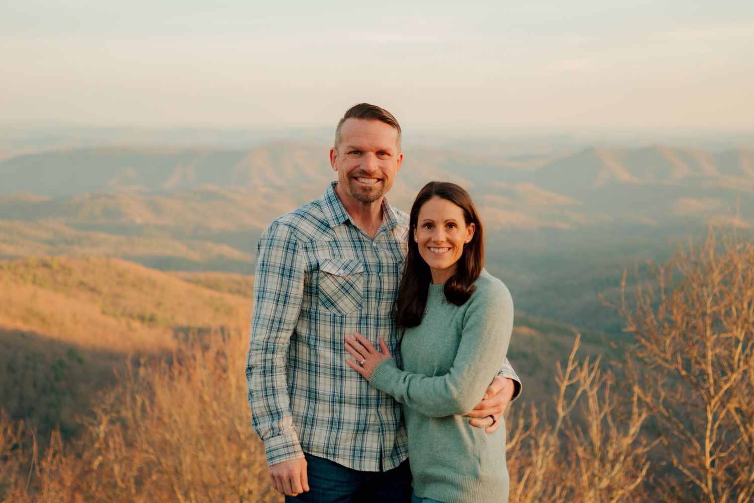 A happy couple embraces on a scenic mountain overlook during golden hour, wearing coordinating blue outfits.