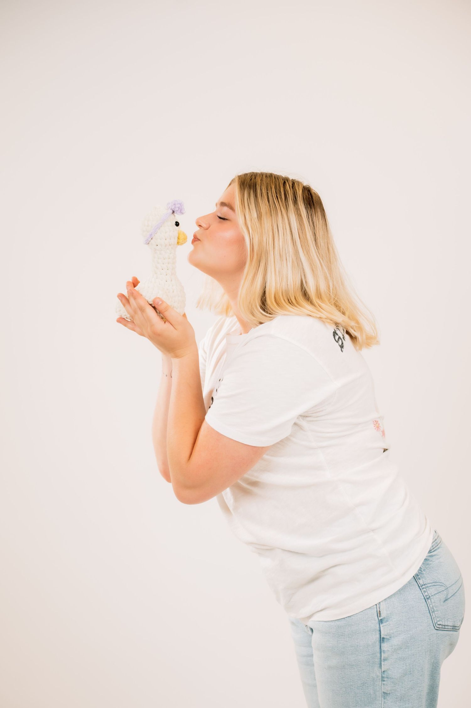 Blonde woman in white tee kissing a small duck stuffed animal toy against white studio background.