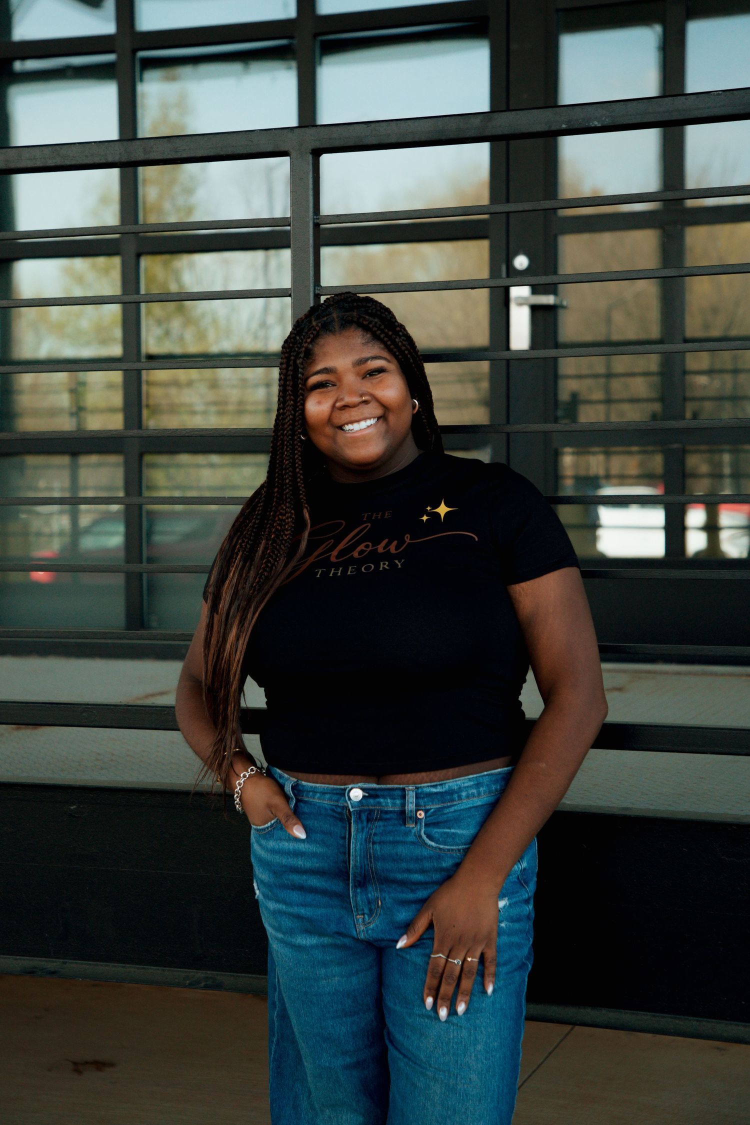 Smiling young woman in black Willow Theory crop top and jeans standing in front of glass windows.