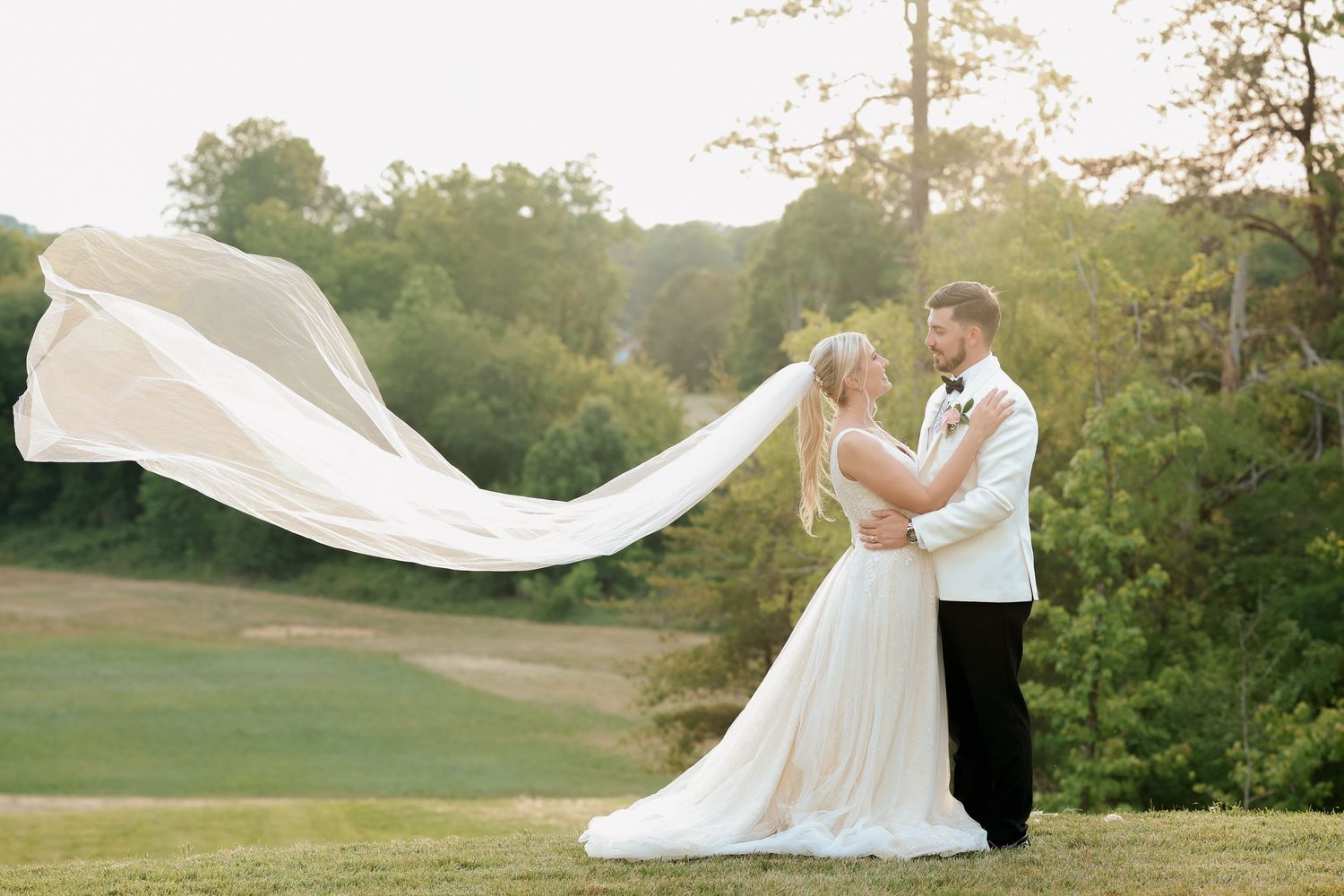 Bride and groom embrace on green hillside with long veil flowing in the breeze.