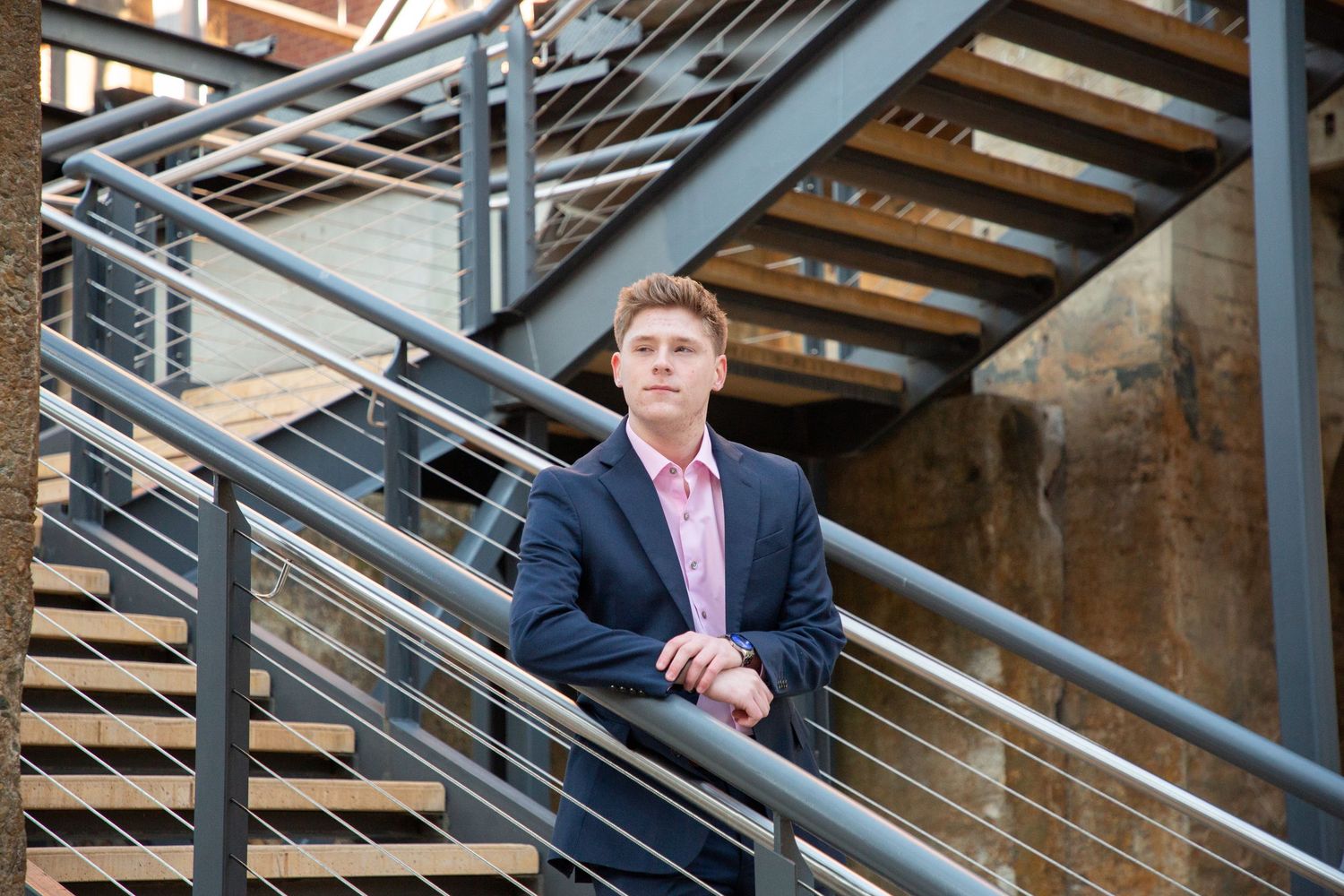 Young man in blue blazer and pink shirt leans on metal staircase railing in urban setting.