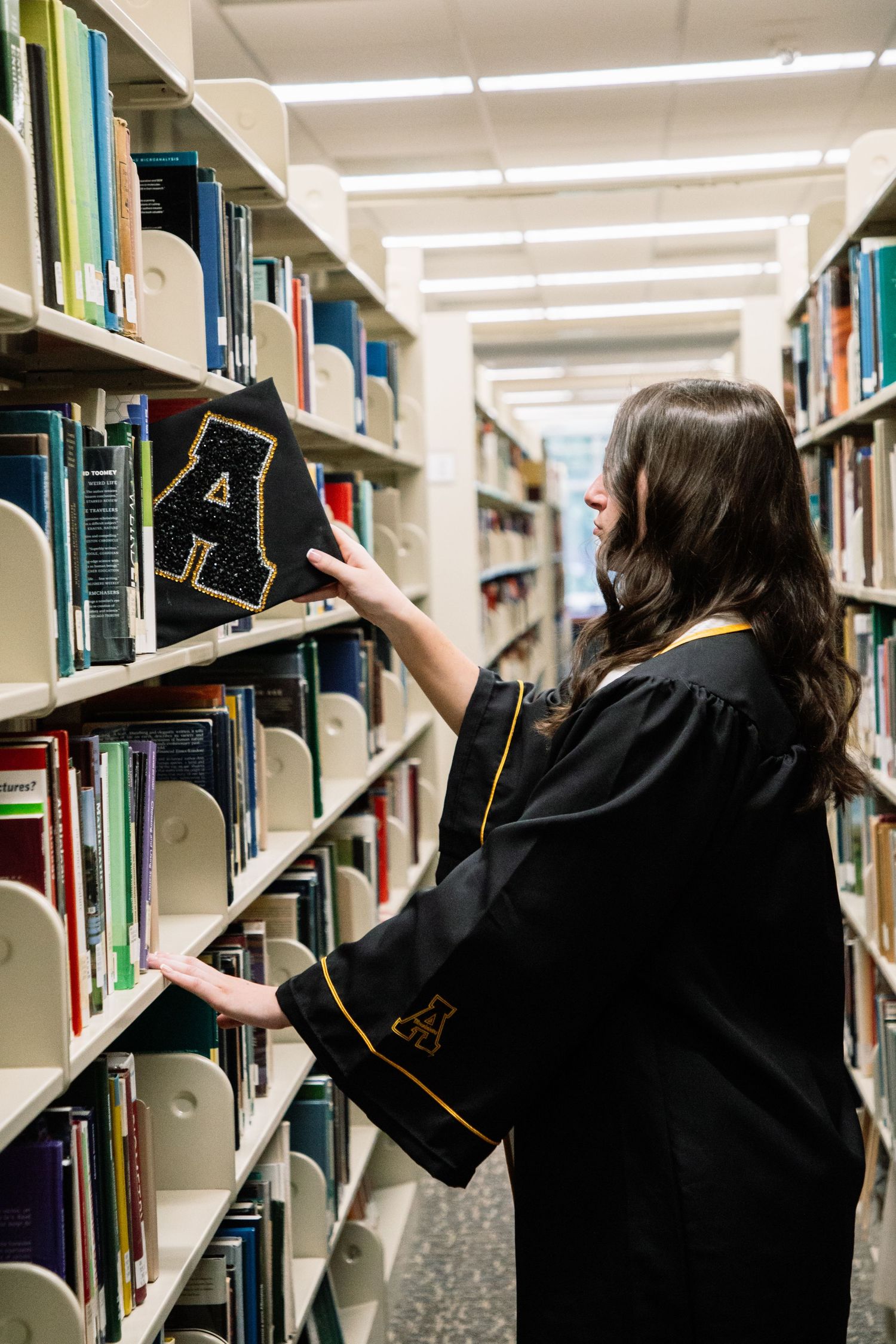 Graduate in black robe placing cap on library shelf among colorful books.