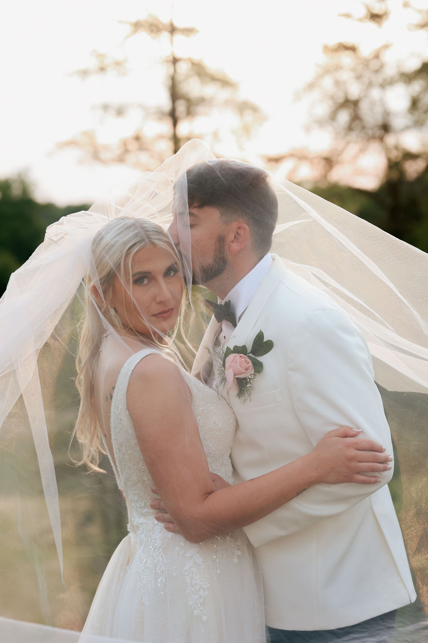 Bride looking at camera while groom kisses her cheek beneath a sheer veil during sunset portraits.