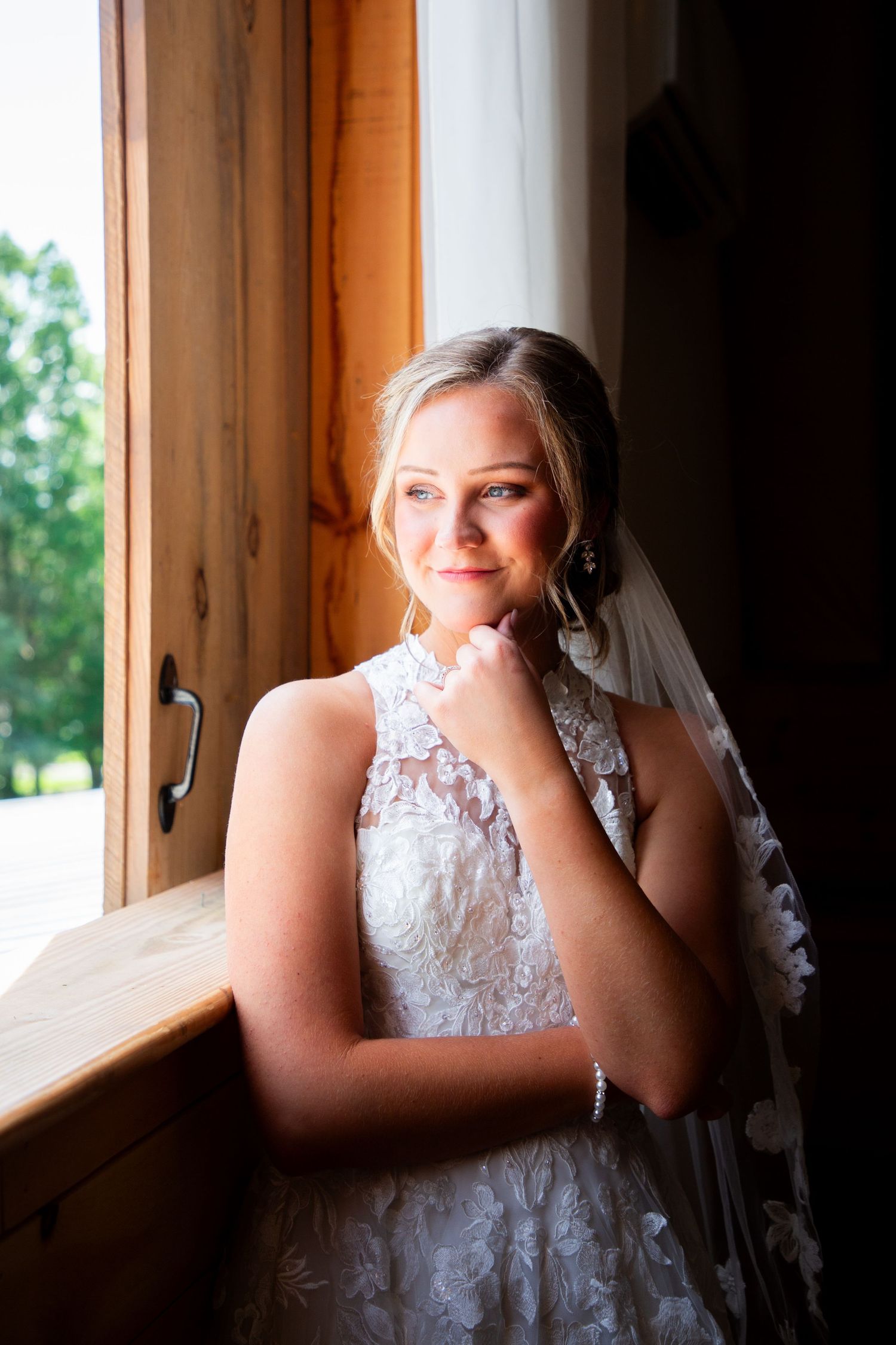 Bride in lace halter gown smiles softly by sunlit rustic wooden window on her wedding day.