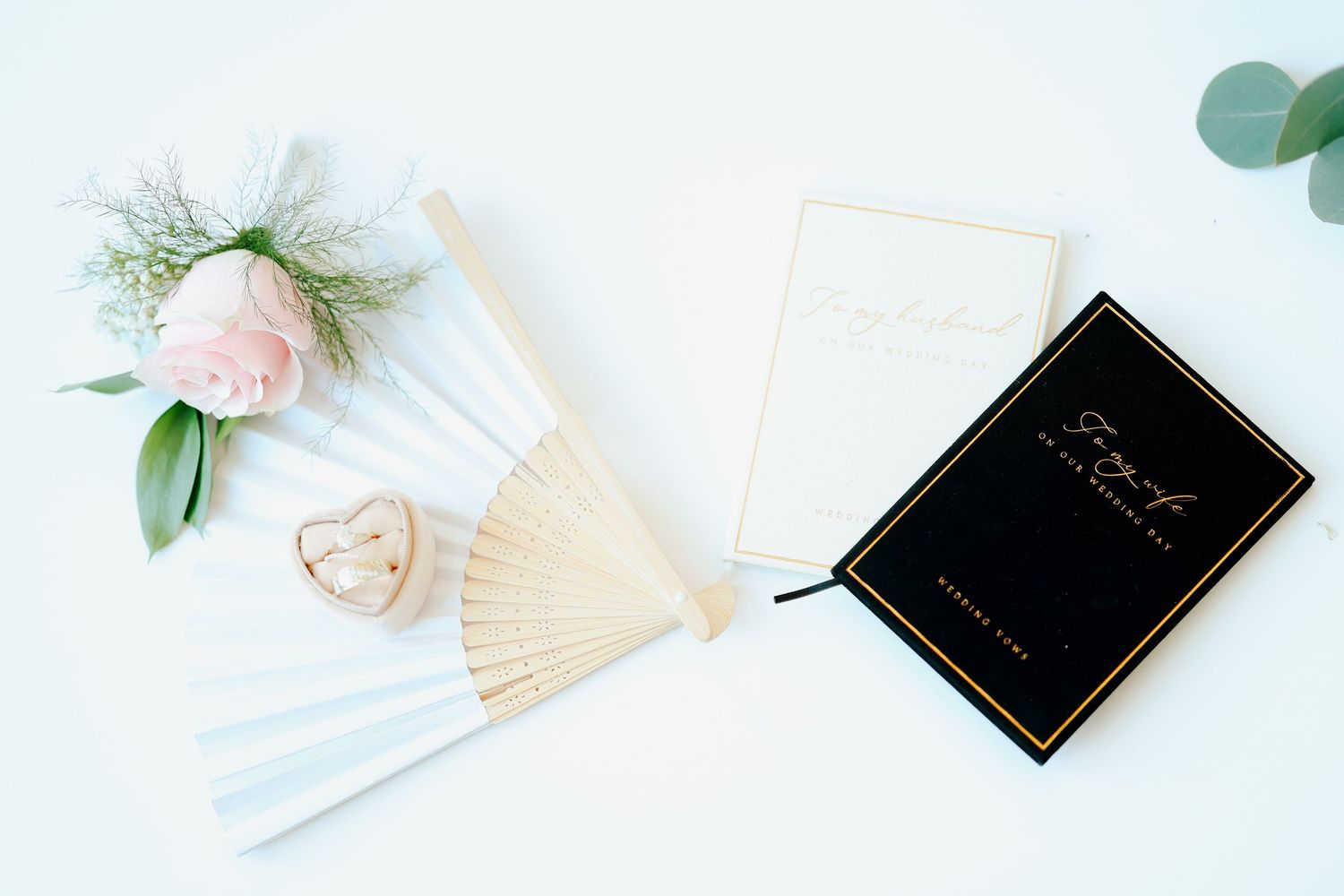 Wedding flat lay with white fan, boutonniere, ring box, and black vow book on white surface.