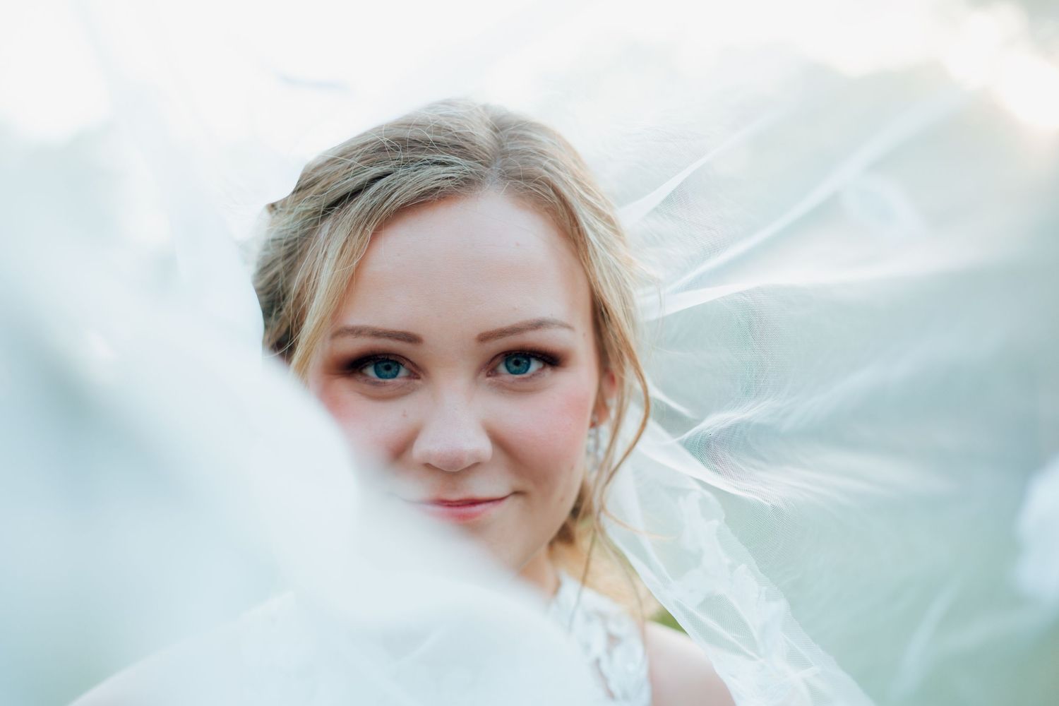 Close-up portrait of blonde bride with blue eyes partially covered by veil.