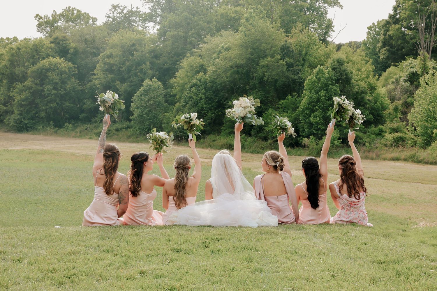 Bride and bridesmaids in blush dresses sitting on green grass with backs turned, raising bouquets in celebration.