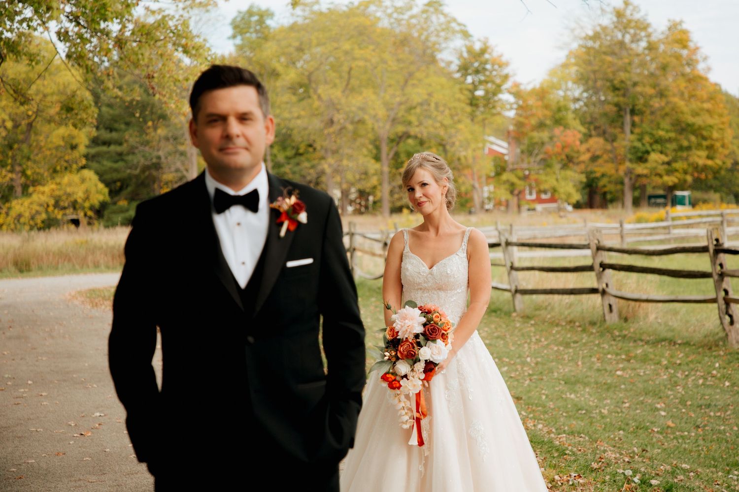 Groom in black tuxedo stands before bride in white lace gown during outdoor fall first look moment.