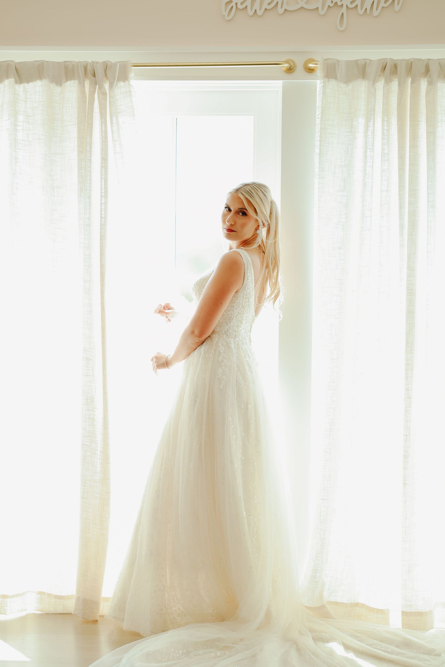 Elegant blonde bride in a flowing white gown standing by a sunlit window.