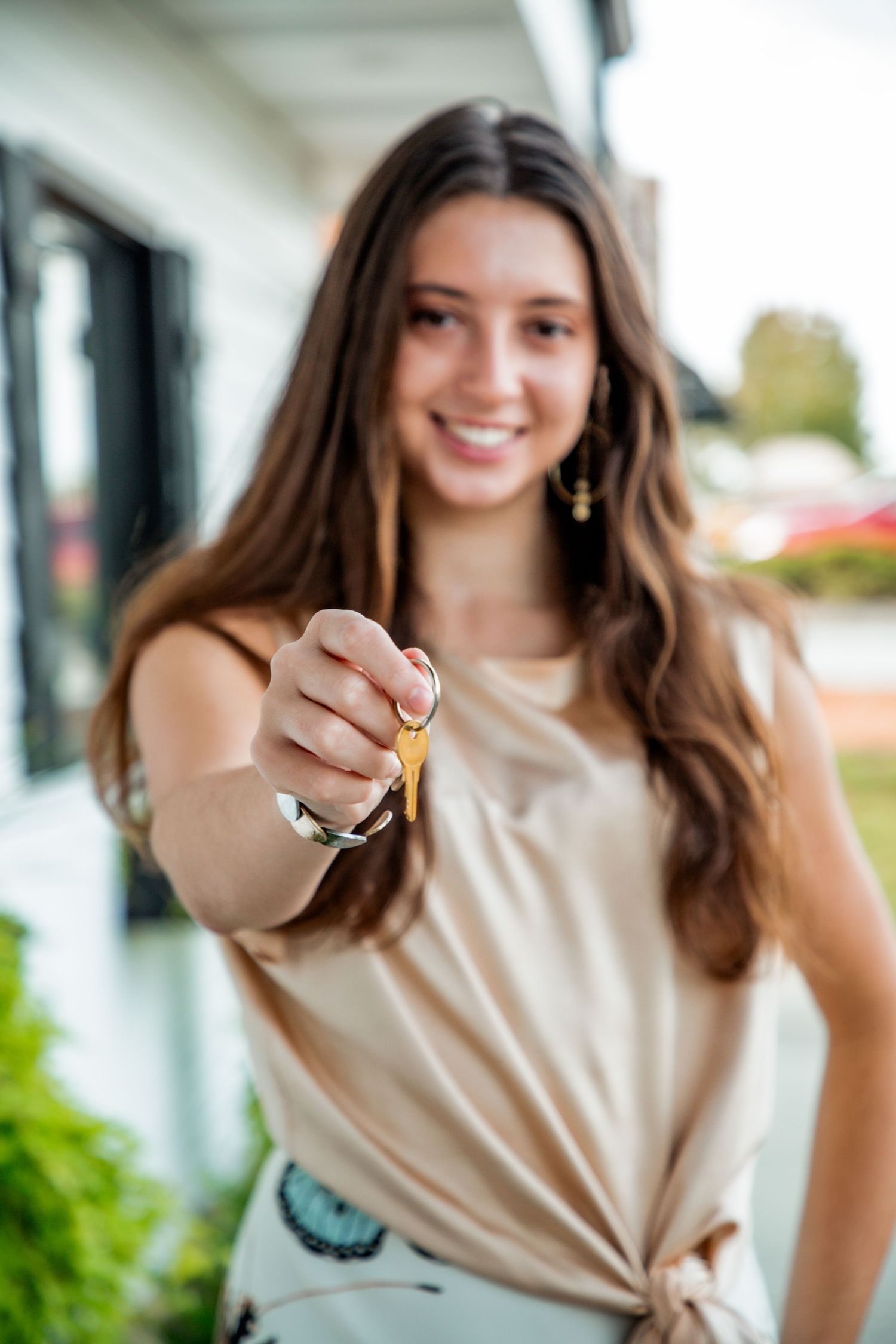 Smiling young woman in beige top holding out house keys toward camera outside new home.