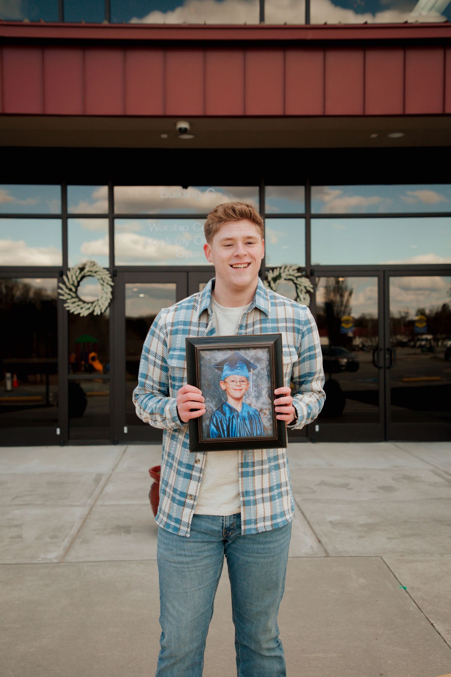 Smiling young man in plaid shirt holding framed graduation photo in front of school.
