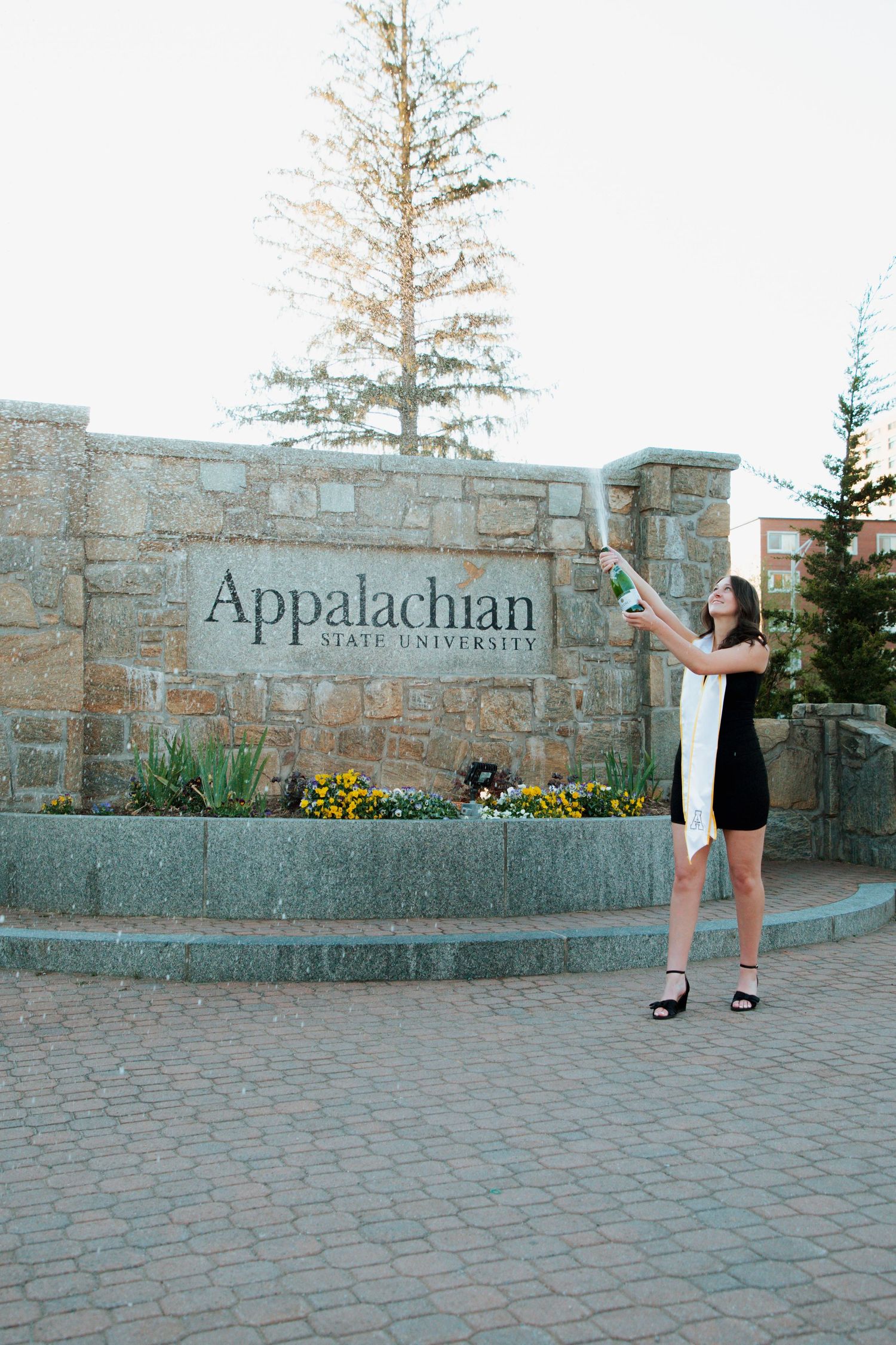 Graduate in black dress pops champagne at Appalachian State University entrance sign.
