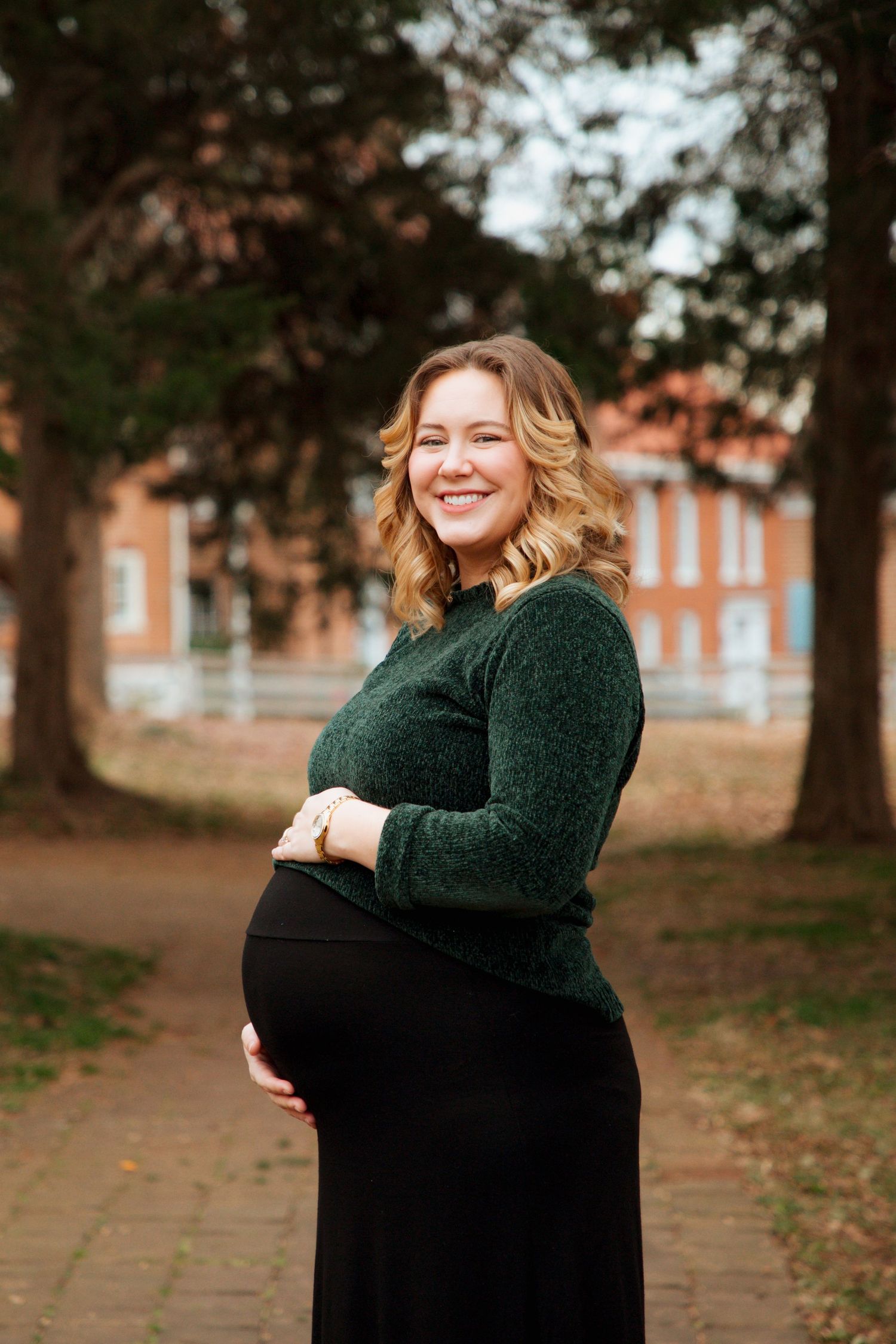 Glowing pregnant woman in dark green sweater and black skirt smiles while cradling baby bump outdoors.