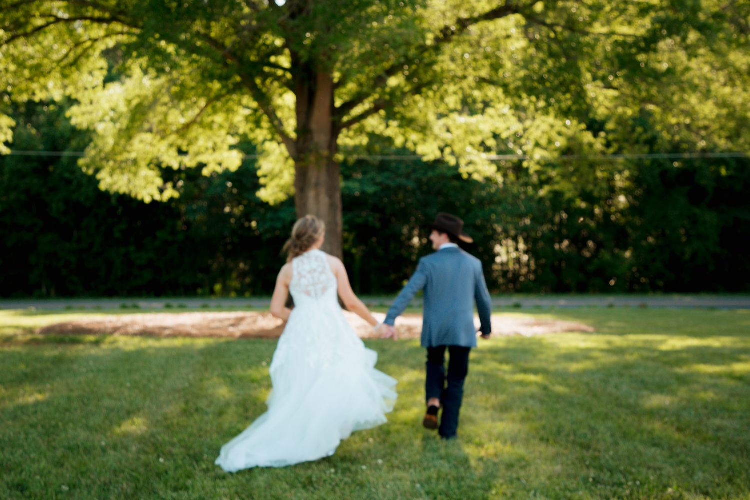 Bride and groom walking hand in hand across lush green lawn toward large tree.